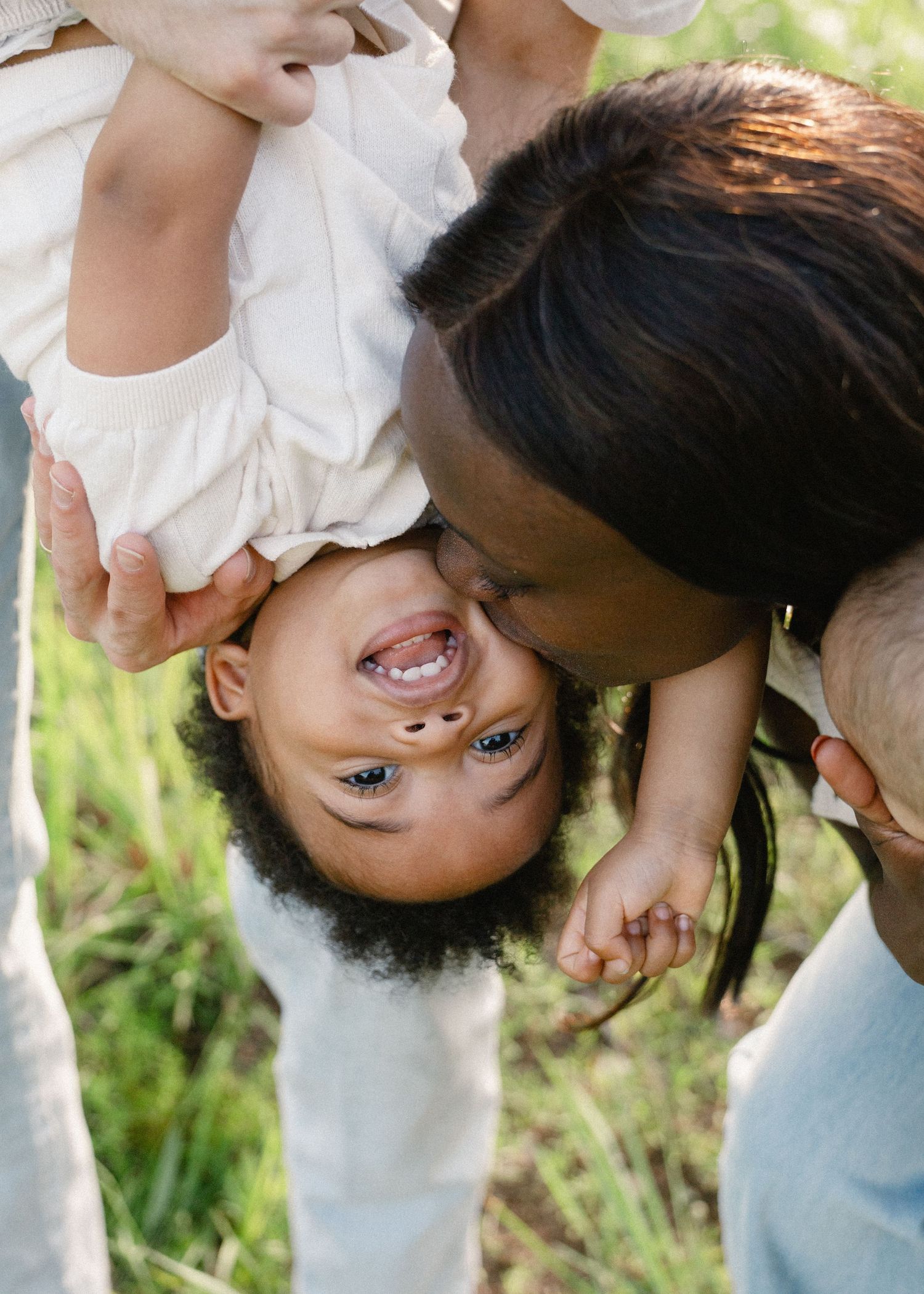 A smiling mother kisses her laughing upside-down toddler in a grassy field.