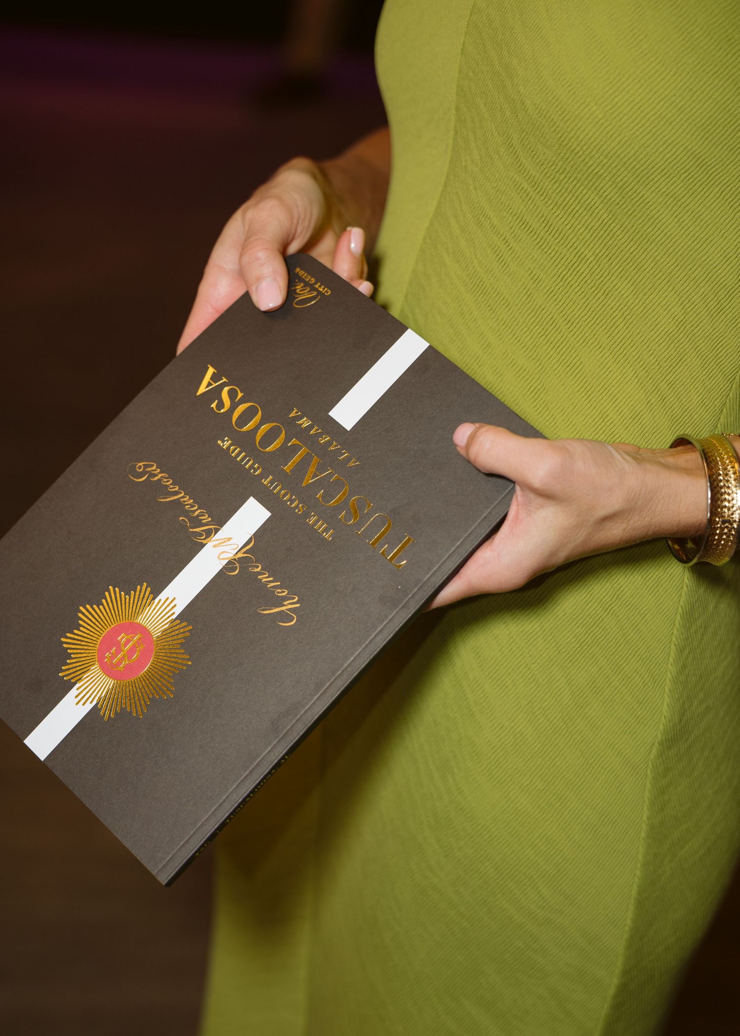 Woman in a lime green dress holding a black Tuscaloosa program booklet with gold foil lettering and a gold medallion emblem.