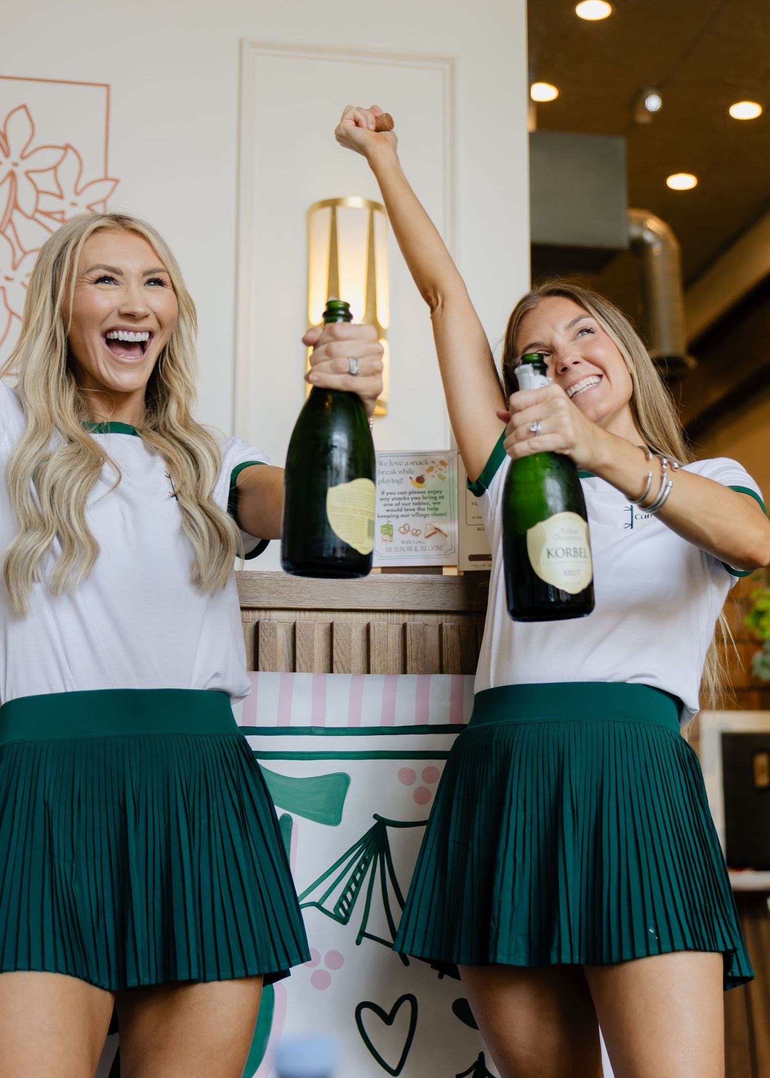 Two women in matching white tops and green skirts celebrate with champagne bottles at a birthday party.