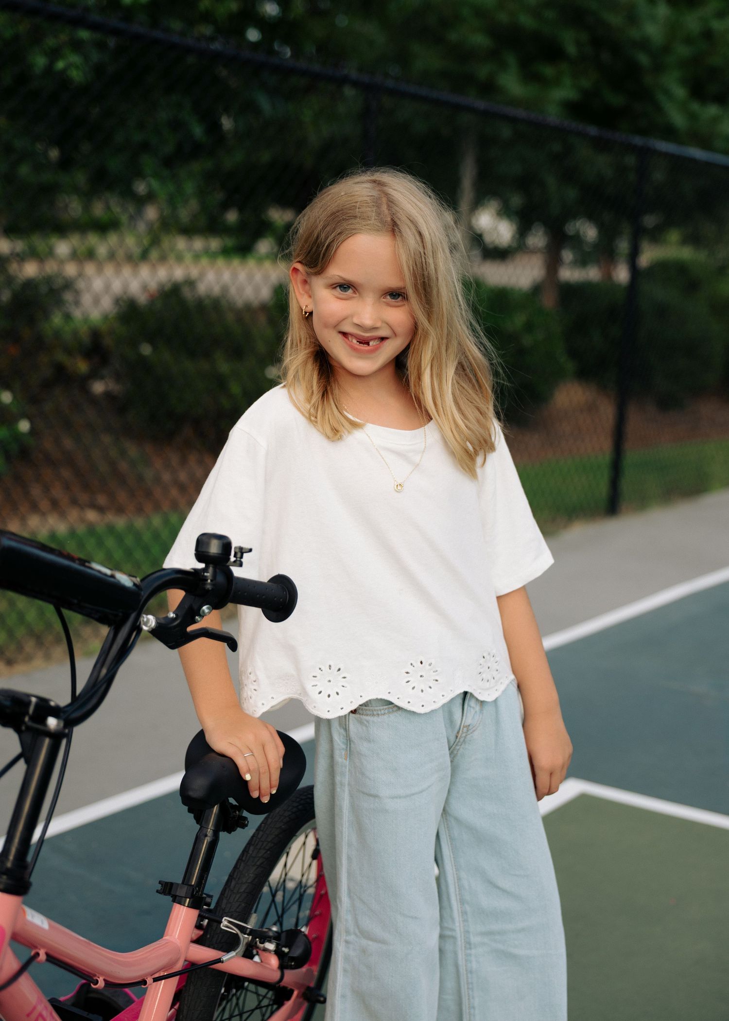 A smiling blonde girl in a white eyelet top stands beside a pink bike on a tennis court.