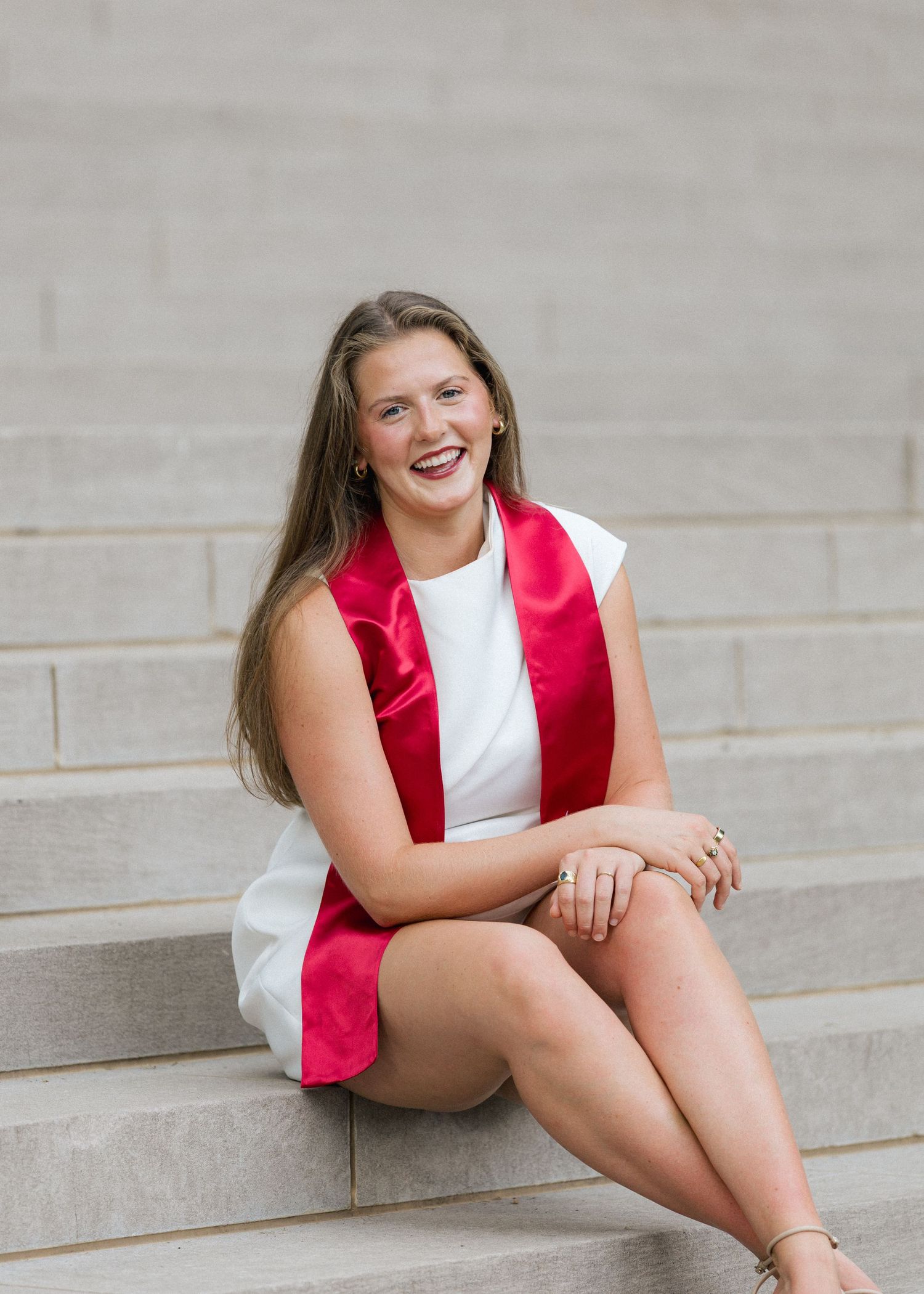 Smiling graduate in white dress and red satin stole sitting on stone steps in outdoor setting.