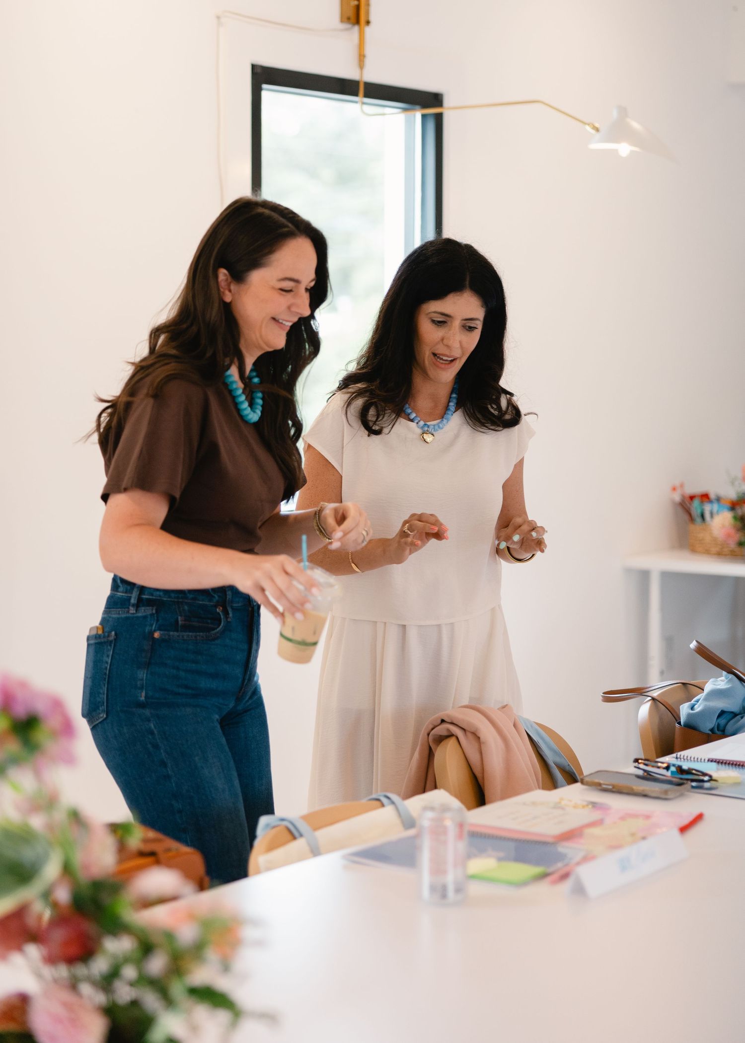 Two women smiling and looking at items on a table in a bright modern studio.