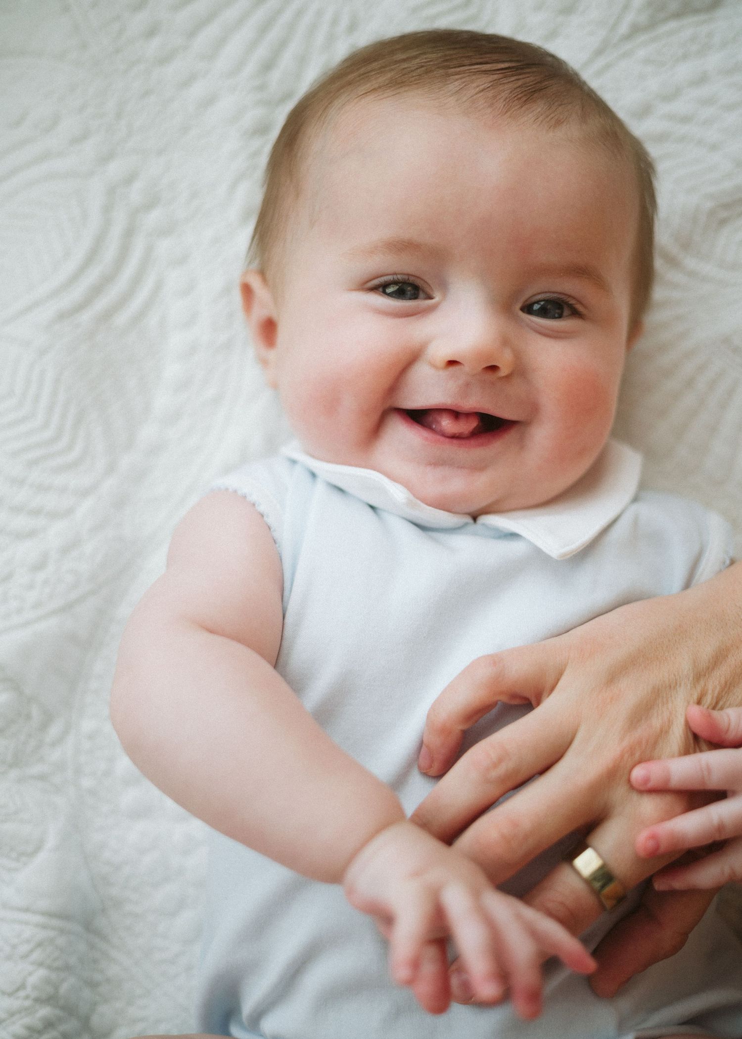 Smiling baby with two teeth lying on a white quilt while an adult holds their hands.