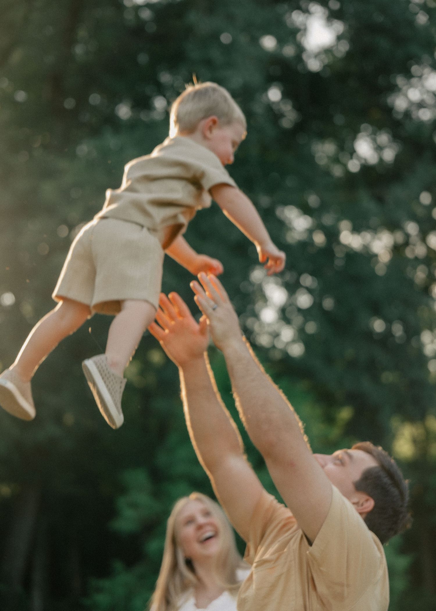 Father tossing laughing toddler in the air while mother watches in sunny park.