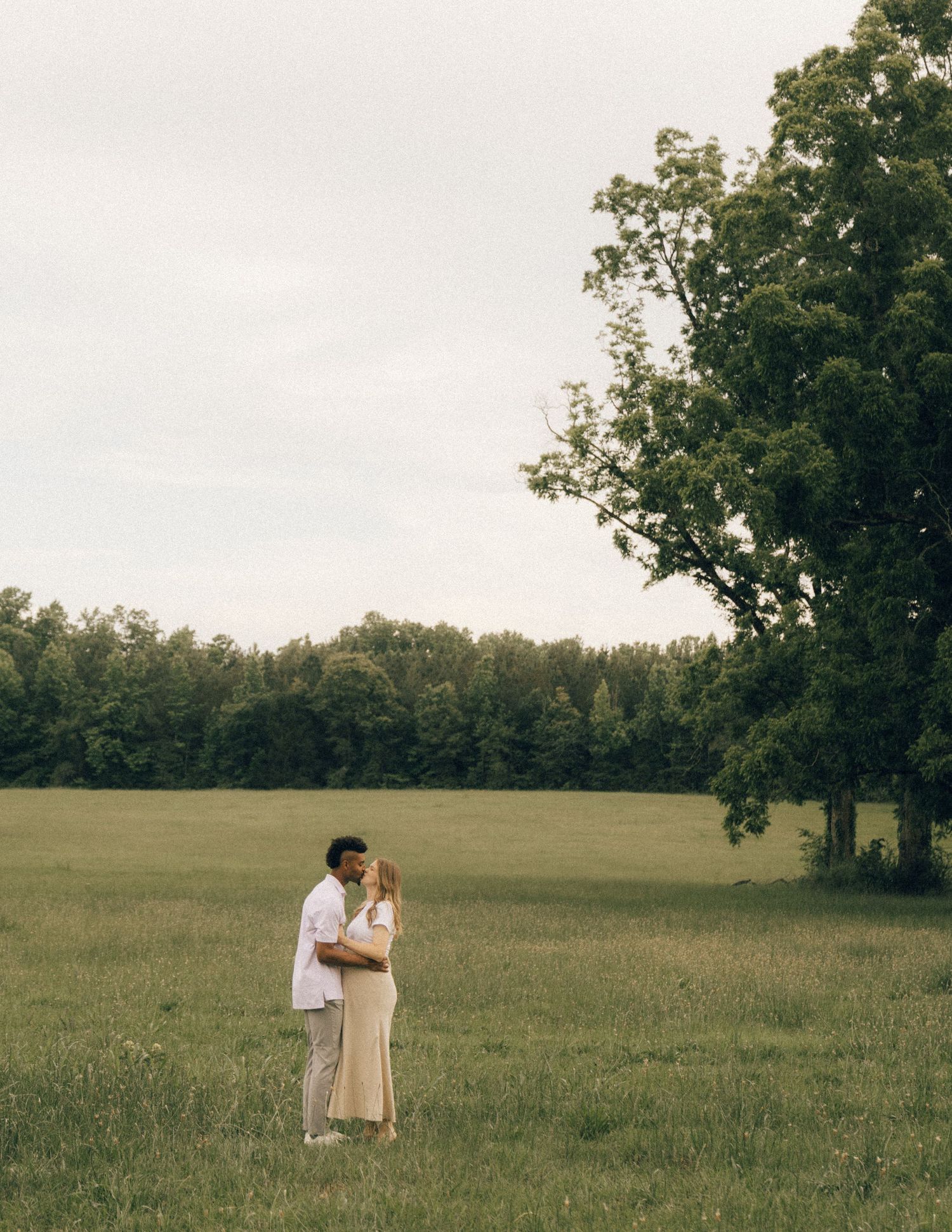 Couple embracing in an open green field with large trees under an overcast sky.