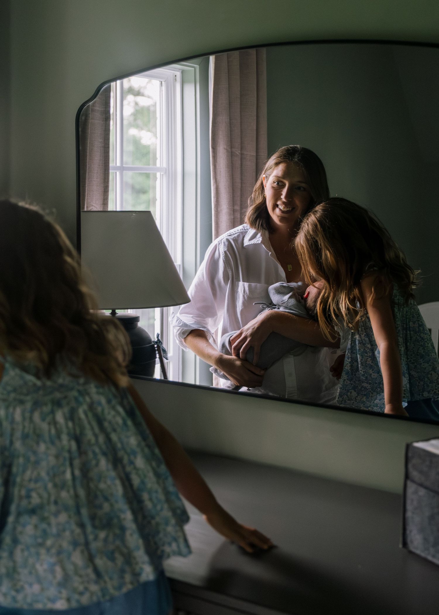 Woman holding newborn baby reflected in large arched mirror with young girl nearby.