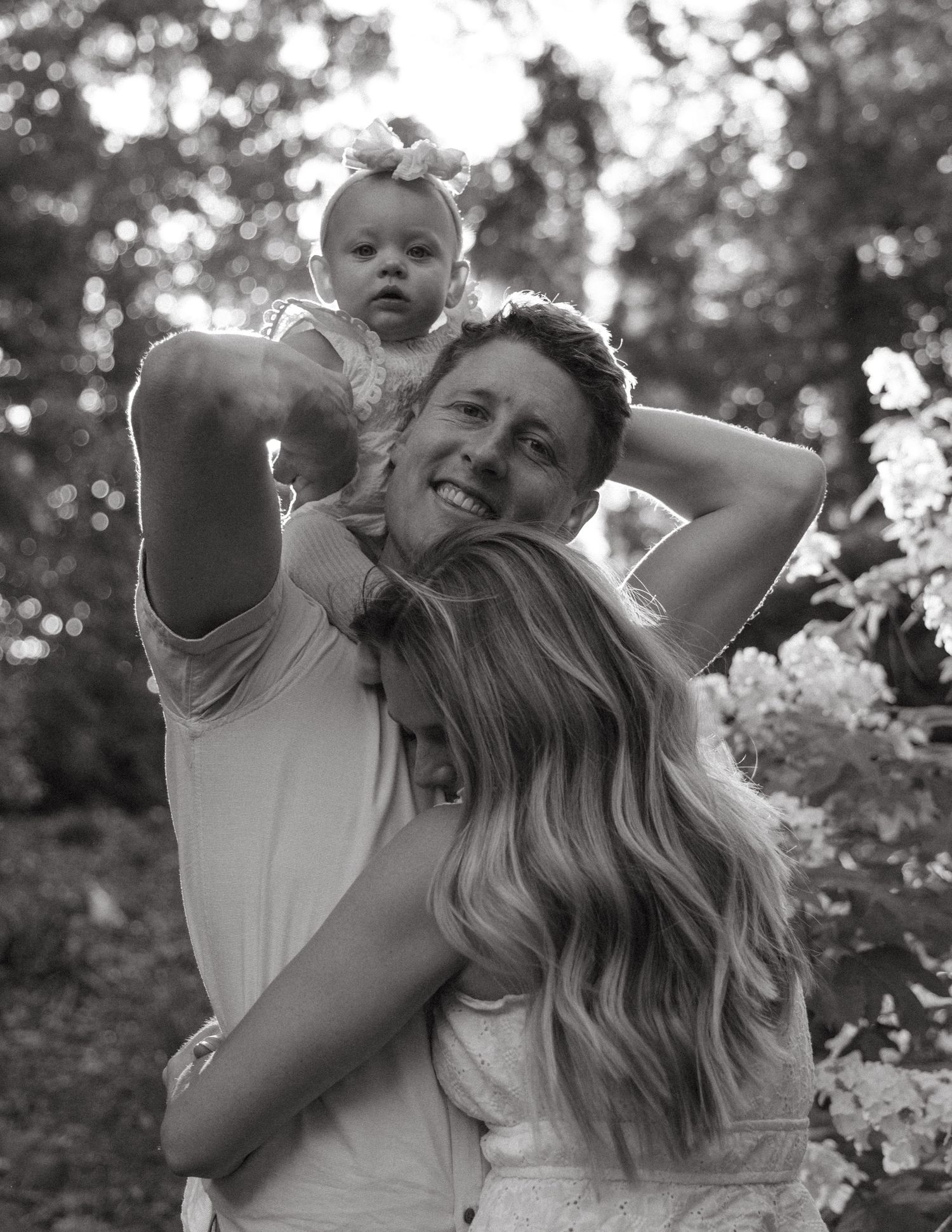 A black and white photo of a couple embracing while their baby sits on dad's shoulders.