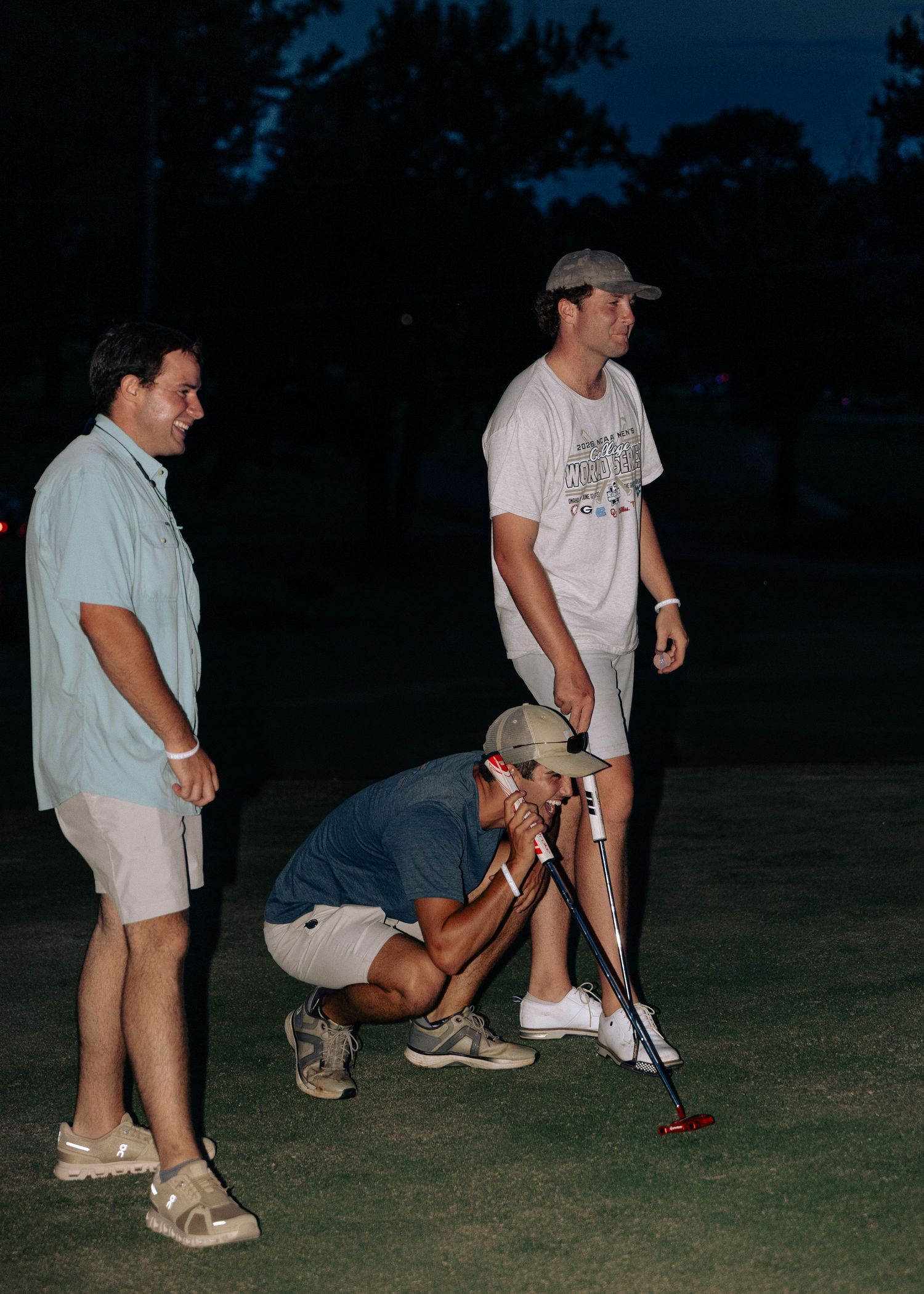Three young men playing mini golf at night on a dimly lit outdoor course.