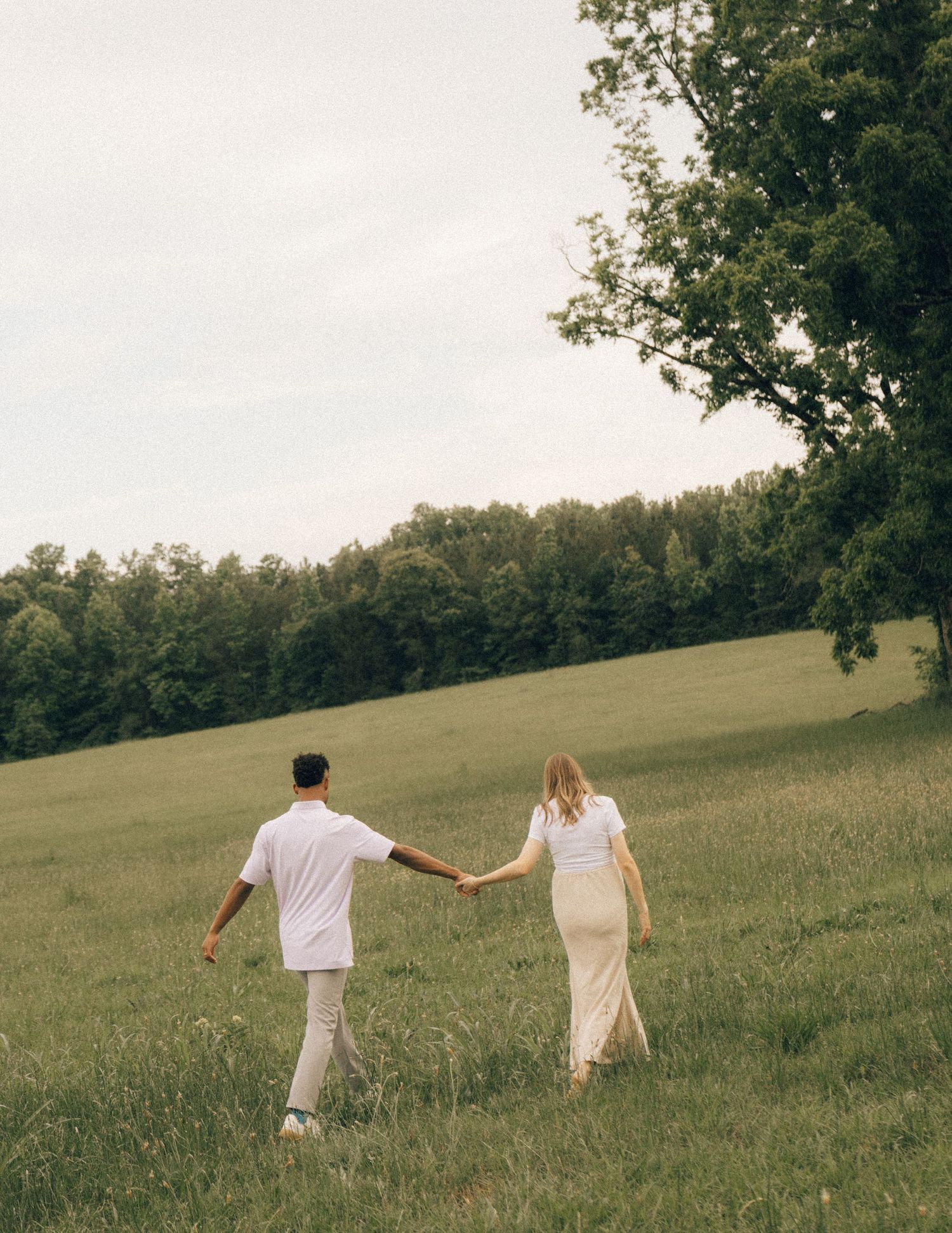 Couple walks hand in hand through a scenic green meadow with tall trees in the background.