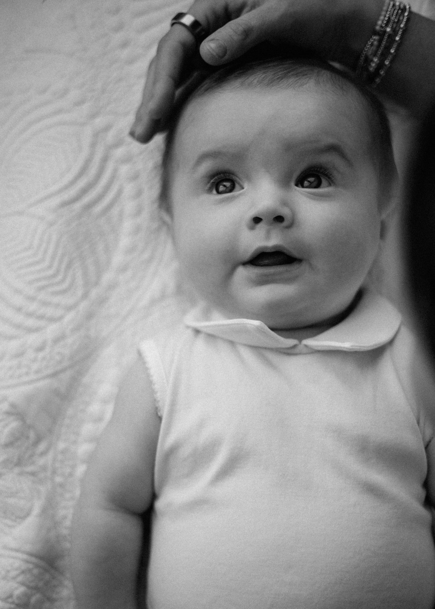 Black and white photo of a curious baby looking up while an adult gently rests a hand on their head.