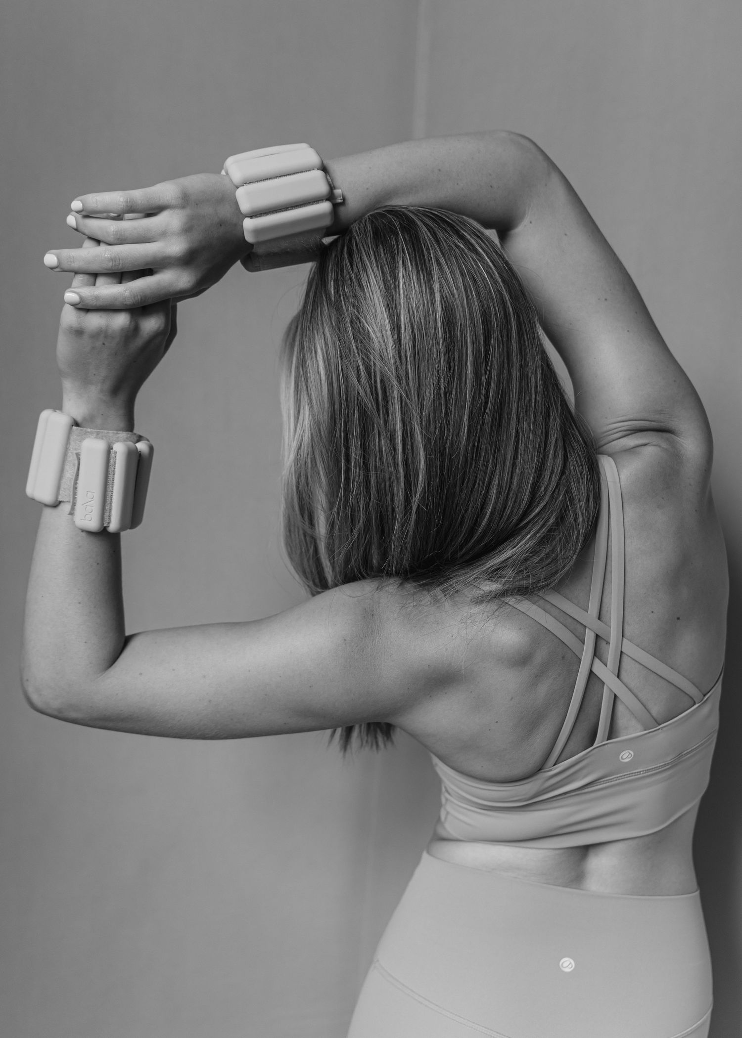 Black and white back view of woman in strappy sports bra with wrist weights raised.