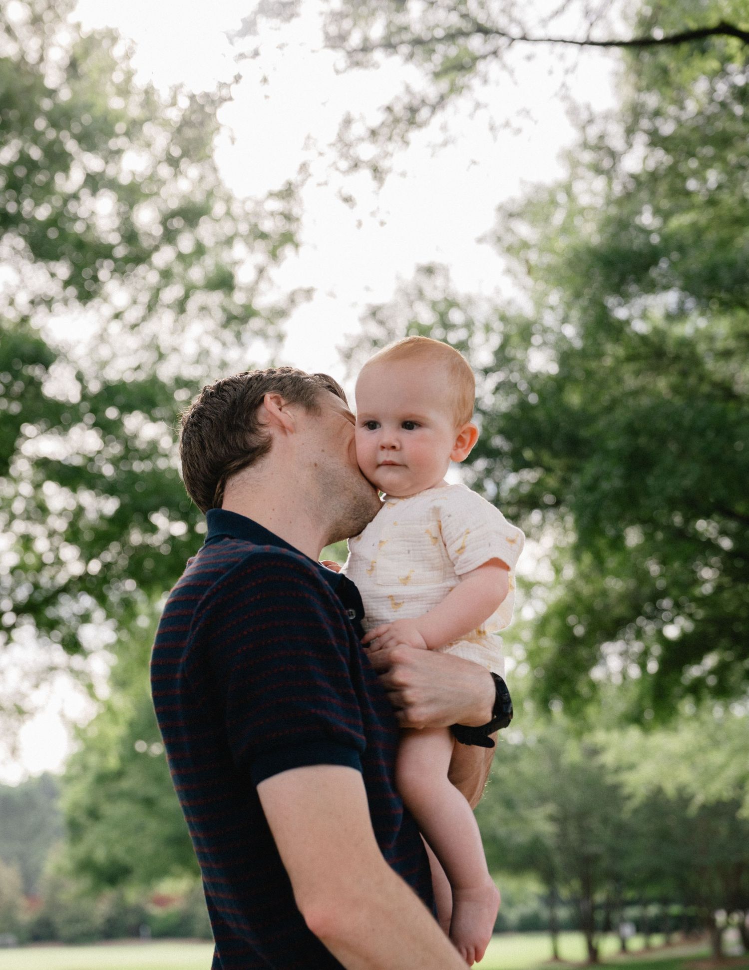 A father tenderly kisses his baby while holding them up in a lush green park.