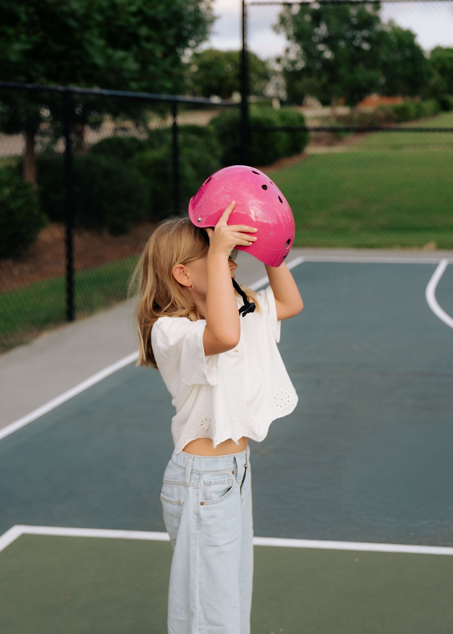 A young blonde girl holds a pink helmet over her face on an outdoor basketball court.
