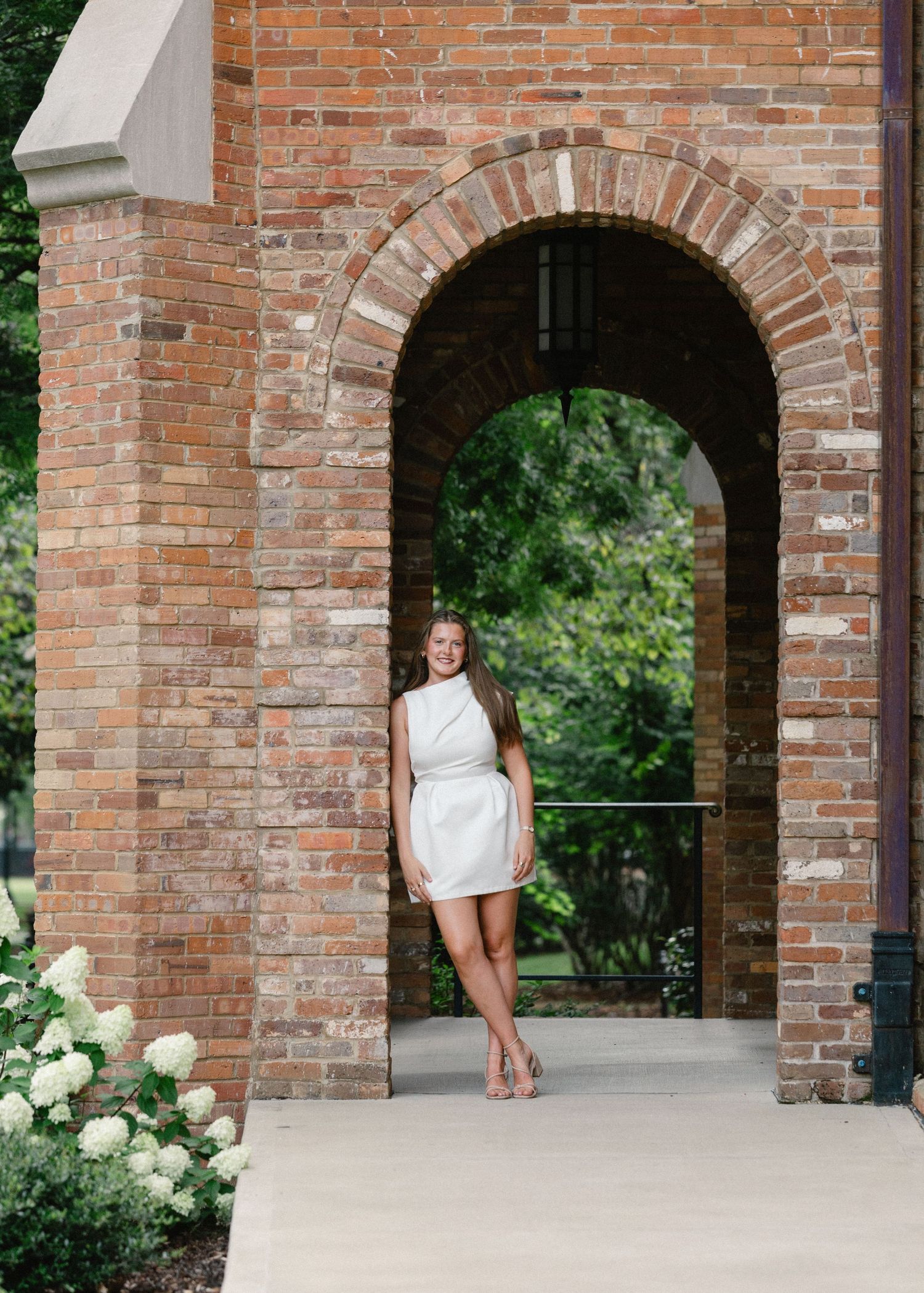 Graduate in white sleeveless dress posing under brick archway with white hydrangeas nearby.