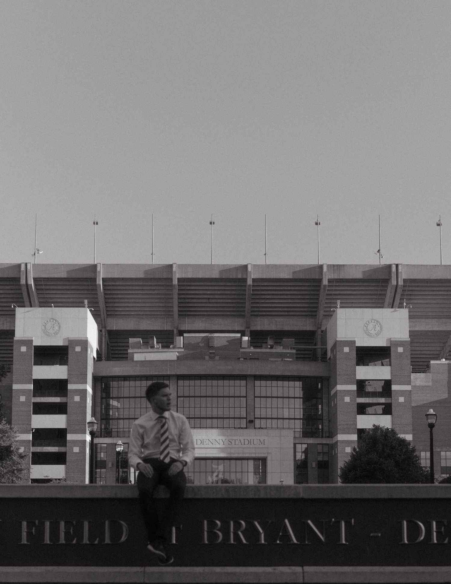Man in dress shirt and tie sitting on wall in front of Bryant-Denny Stadium.