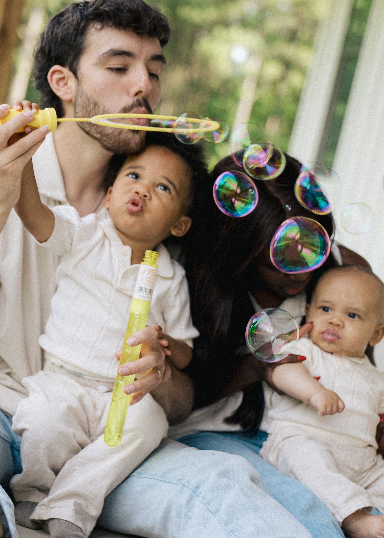 Family blowing bubbles together outdoors, with a toddler and baby joining in the fun.
