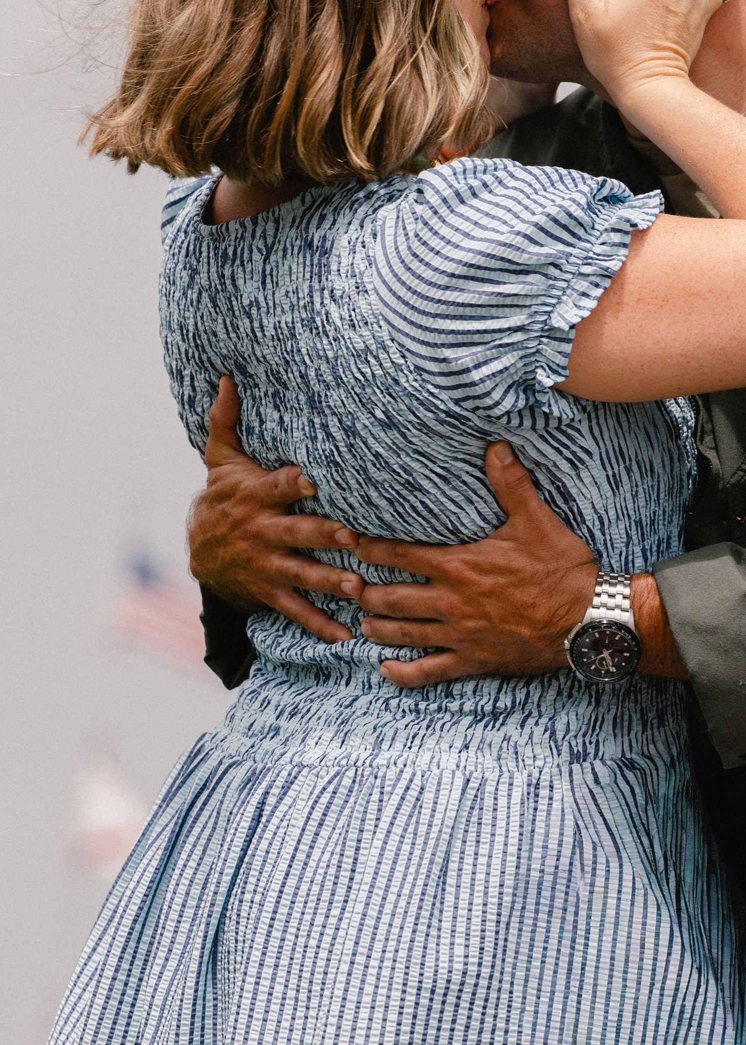A couple shares an emotional embrace, the man's hand resting on the woman's blue striped dress during a military homecoming.