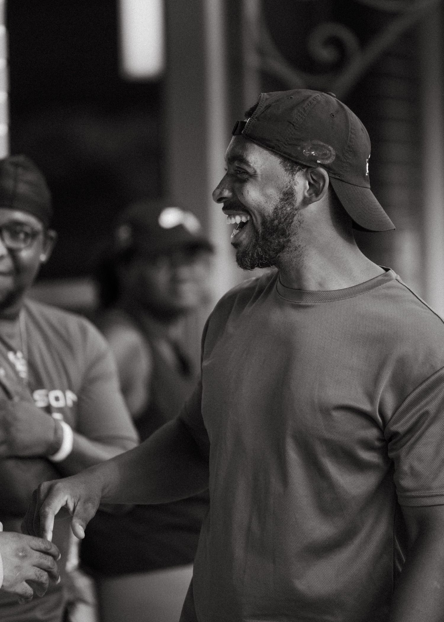 Black and white photo of a laughing man in a baseball cap and t-shirt at an outdoor gathering.