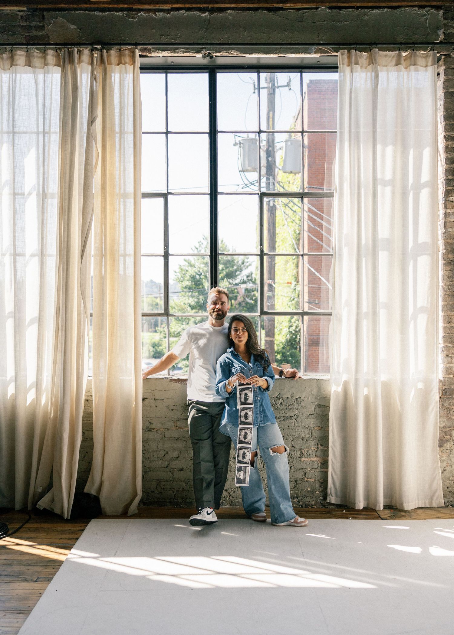 Excited couple standing by large industrial windows holding ultrasound photos in a bright loft studio.