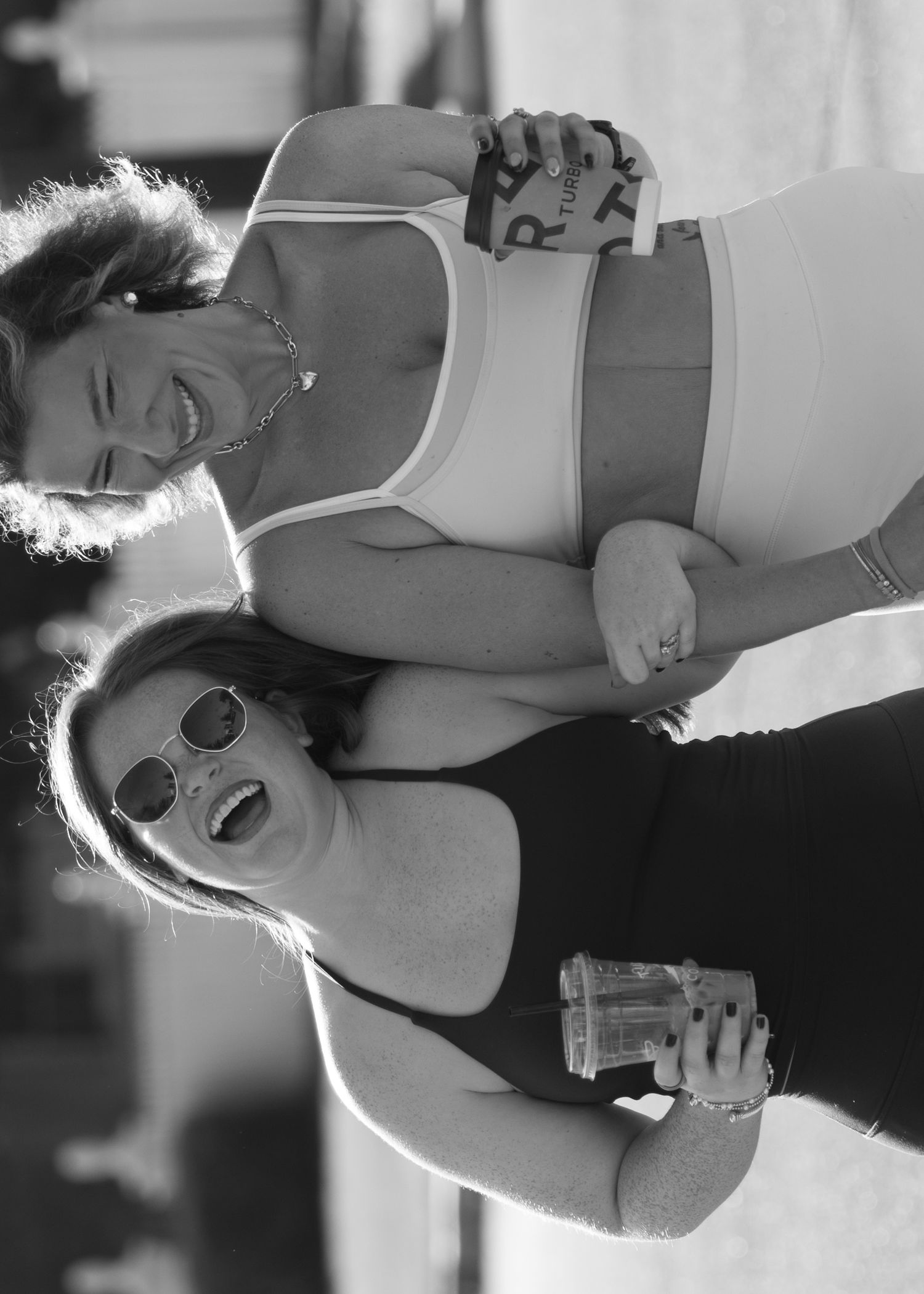 Two women laughing together outdoors holding drinks in black and white candid fitness photo.