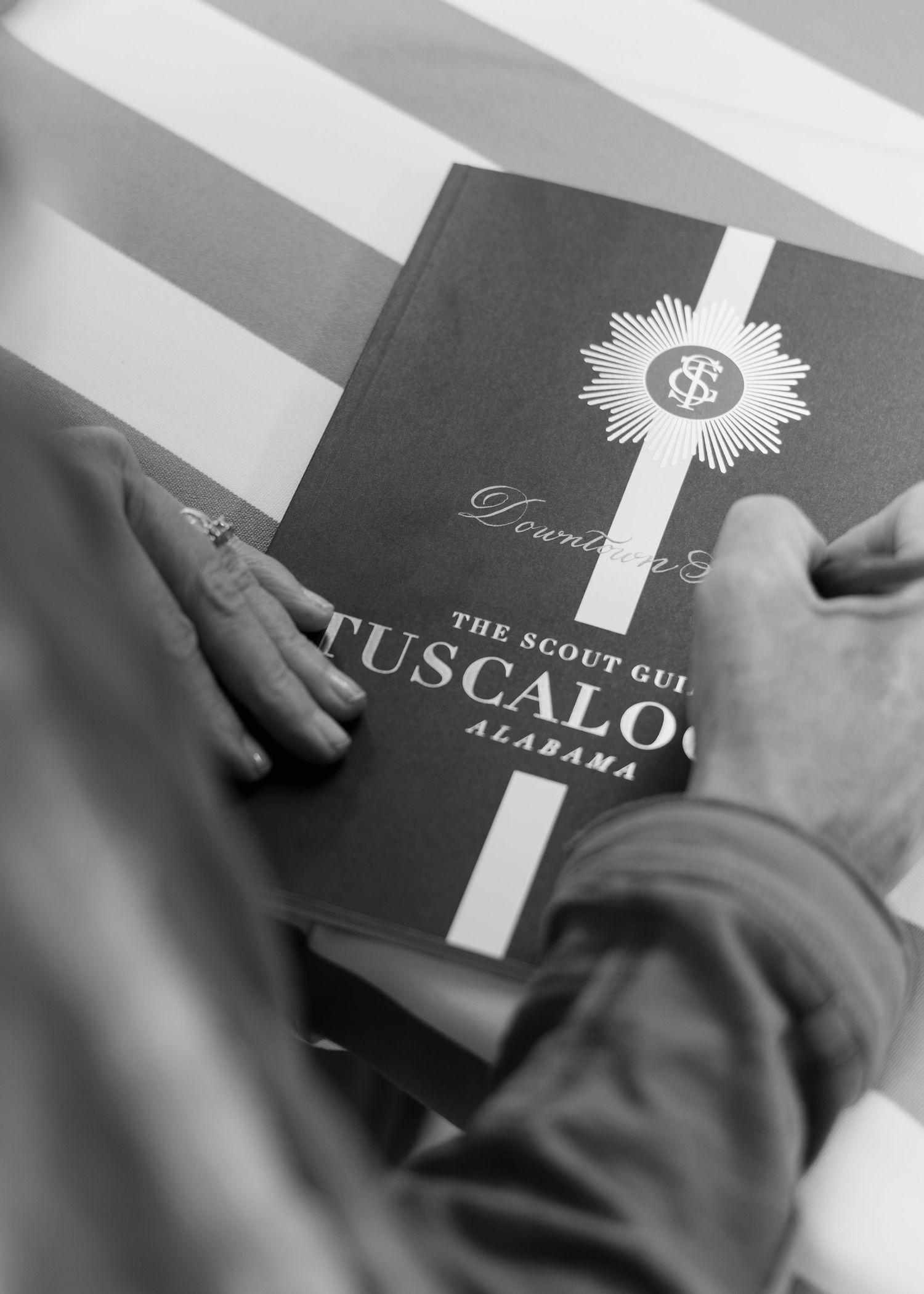 Hands rest on The Scout Guide Tuscaloosa, Alabama, a black and white photo of a book with a sunburst emblem.
