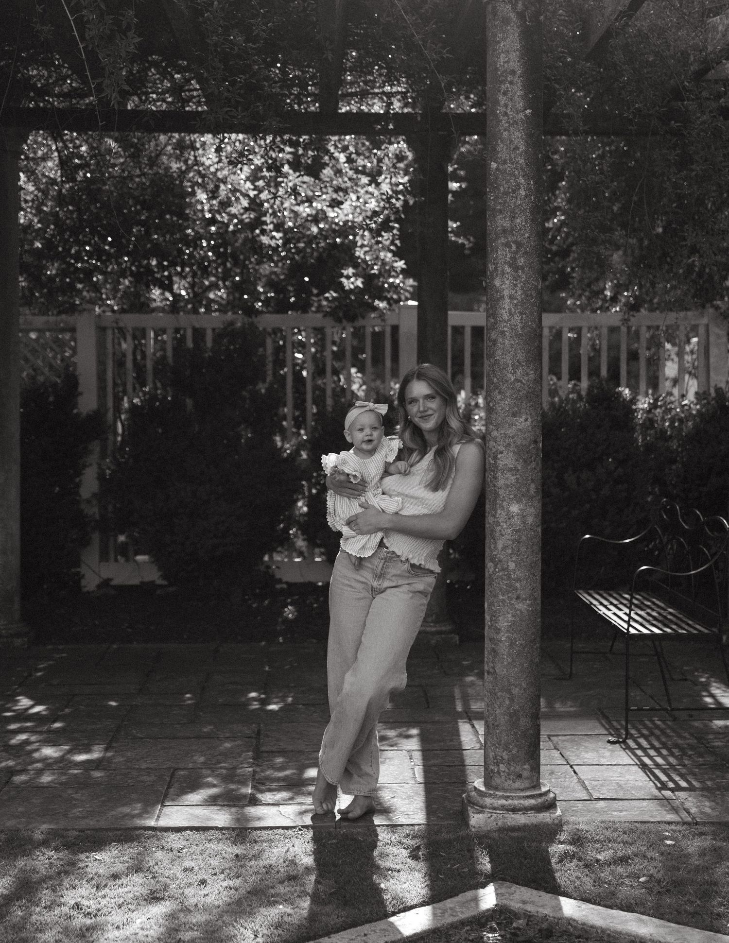 A young mother holds her baby while leaning against a pergola post in black and white.