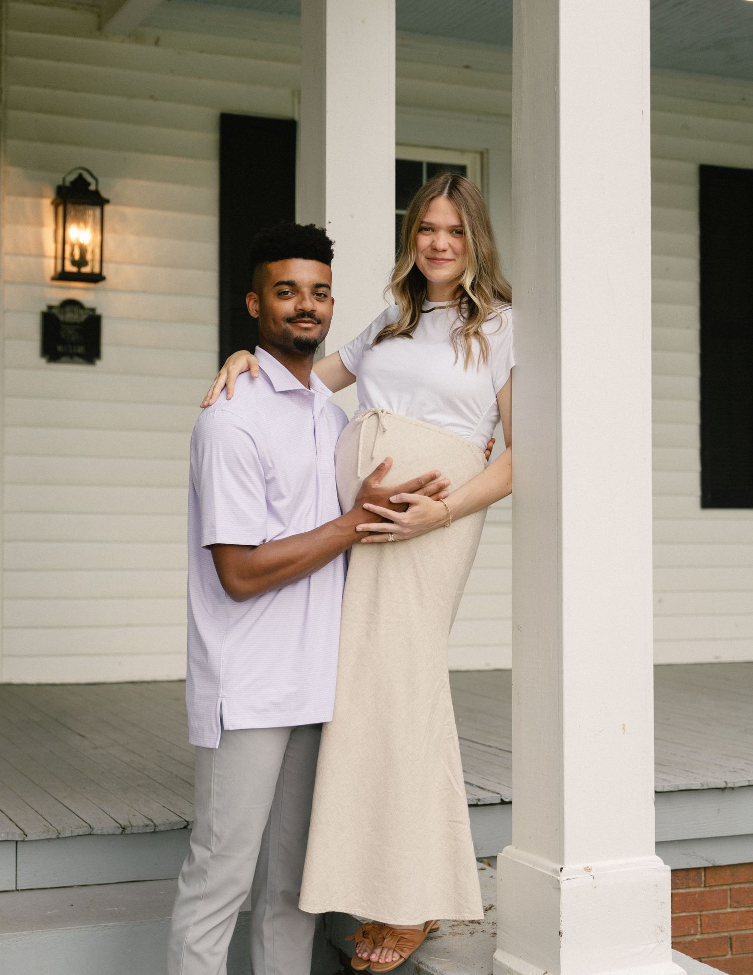 Expecting couple poses on a white farmhouse porch, man in lavender shirt embraces pregnant woman in cream dress.