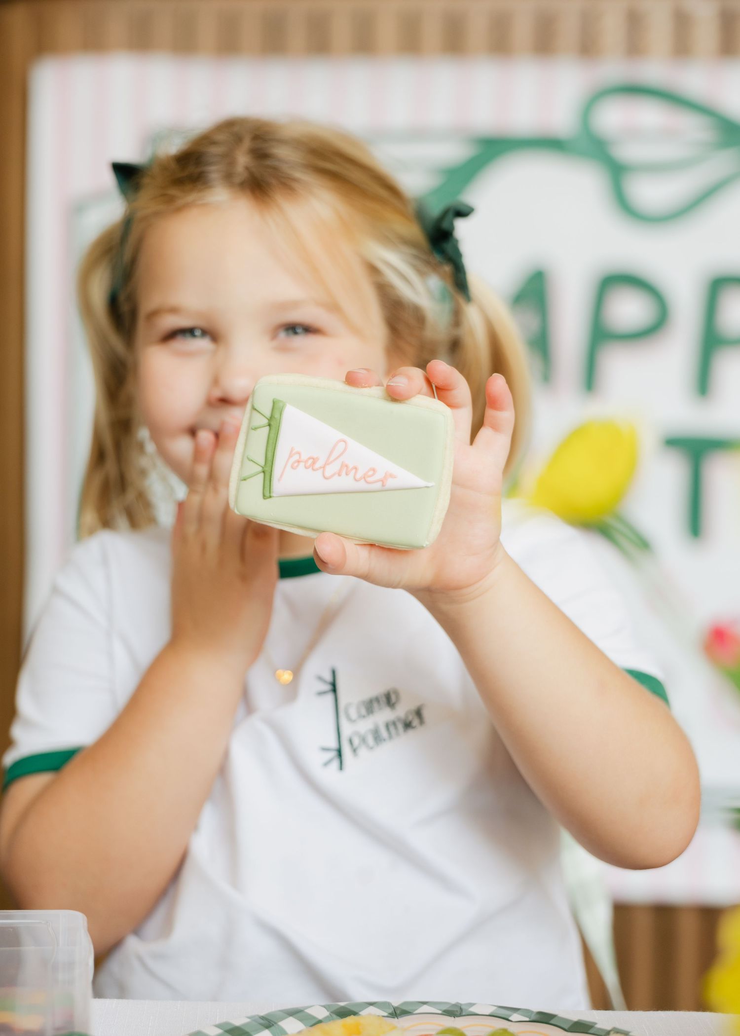Young blonde girl holds up a decorated camp-themed birthday cookie at a birthday party.