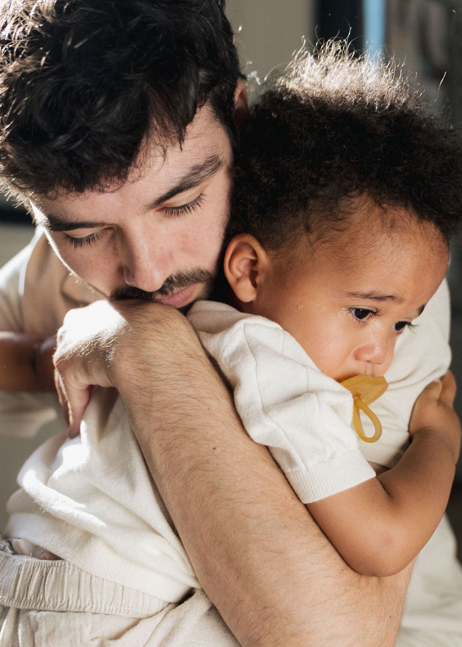 Father tenderly embracing his toddler who holds a pacifier in a close emotional moment.