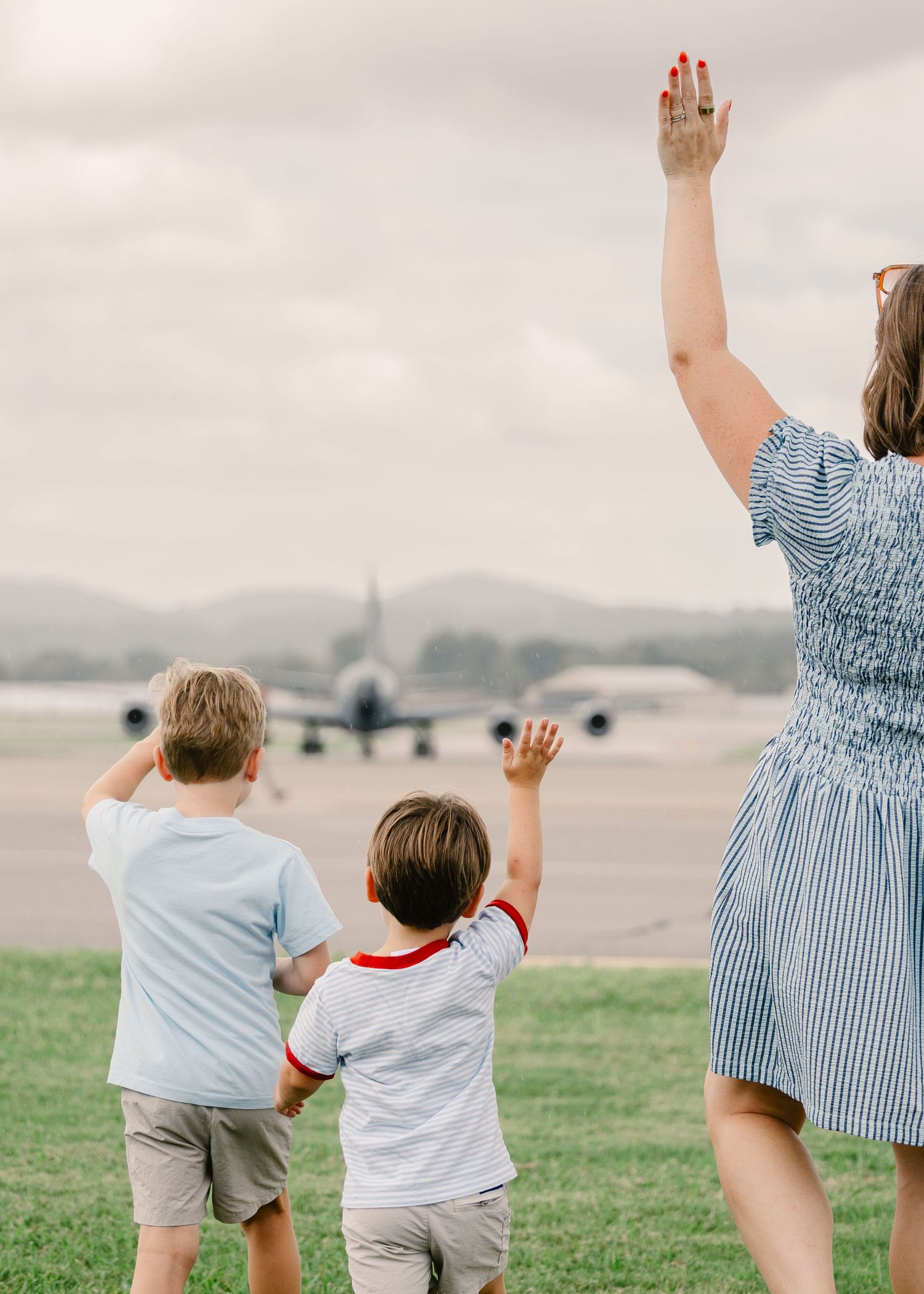 A mother and two young boys wave goodbye to a military aircraft taxiing on the runway from the airfield grass.