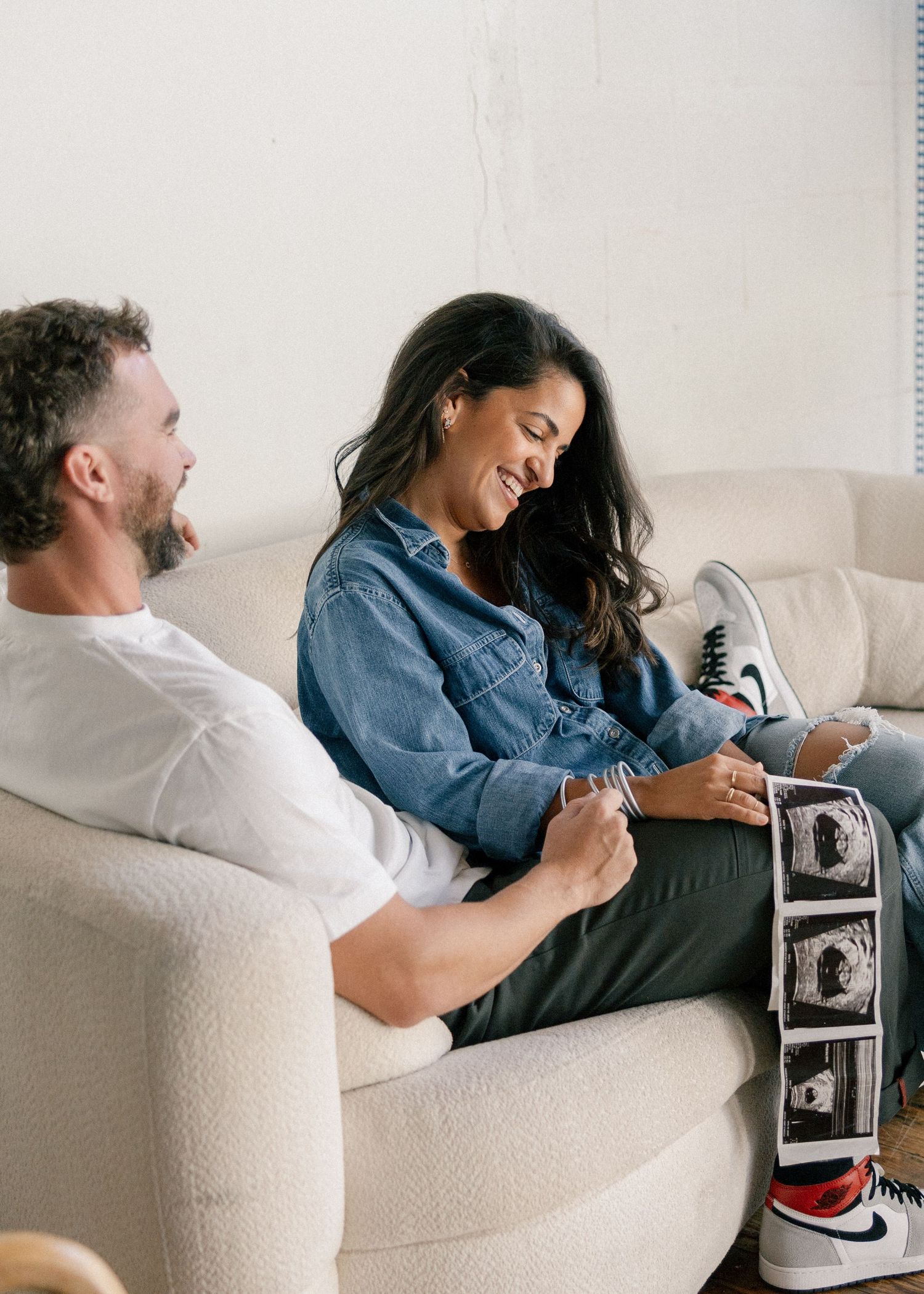 Happy couple sitting on a couch smiling at ultrasound photos during an intimate pregnancy announcement session.