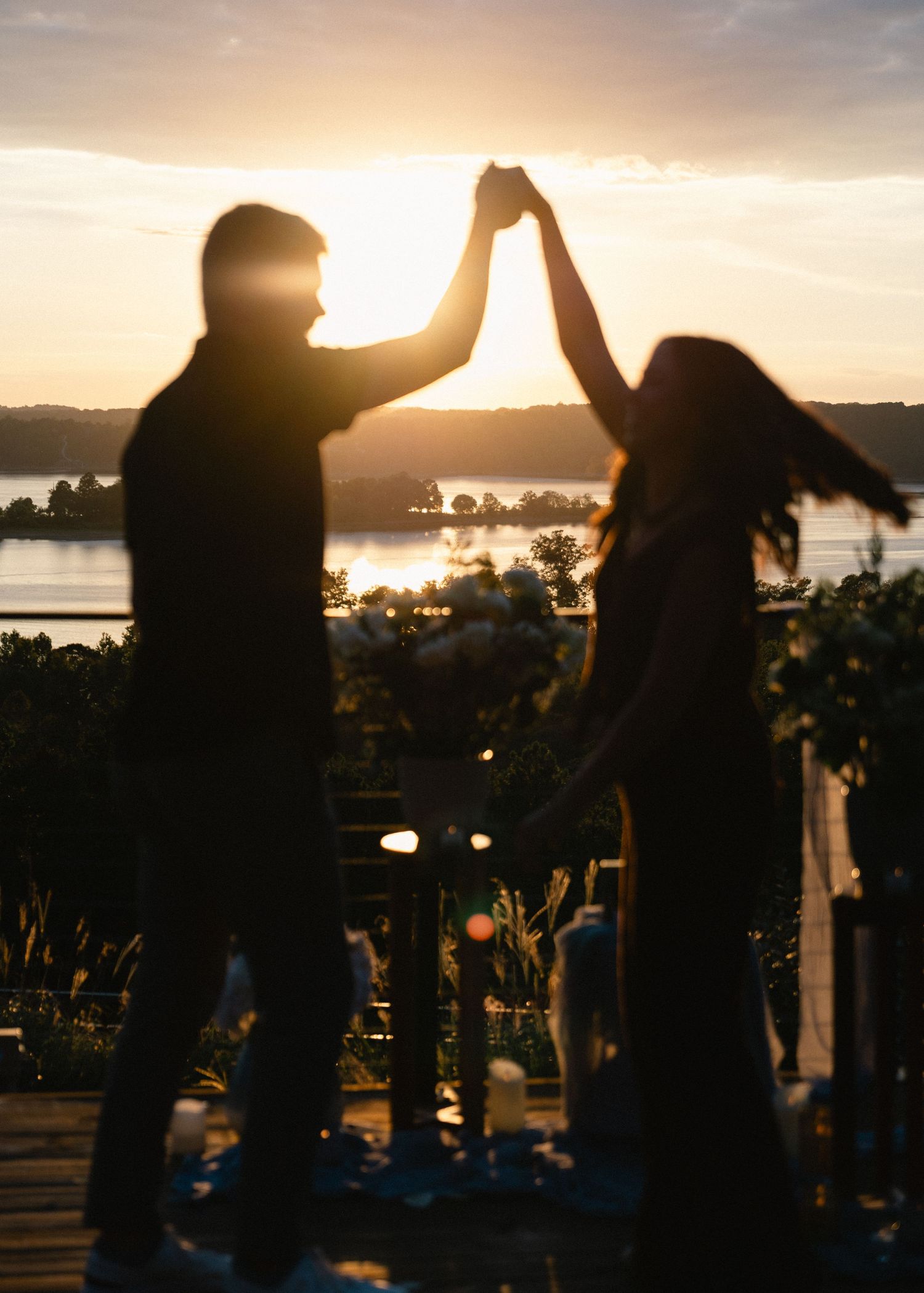 Silhouetted couple dancing hand in hand at golden hour beside a candlelit table with flowers, with a glowing sun over the lake.