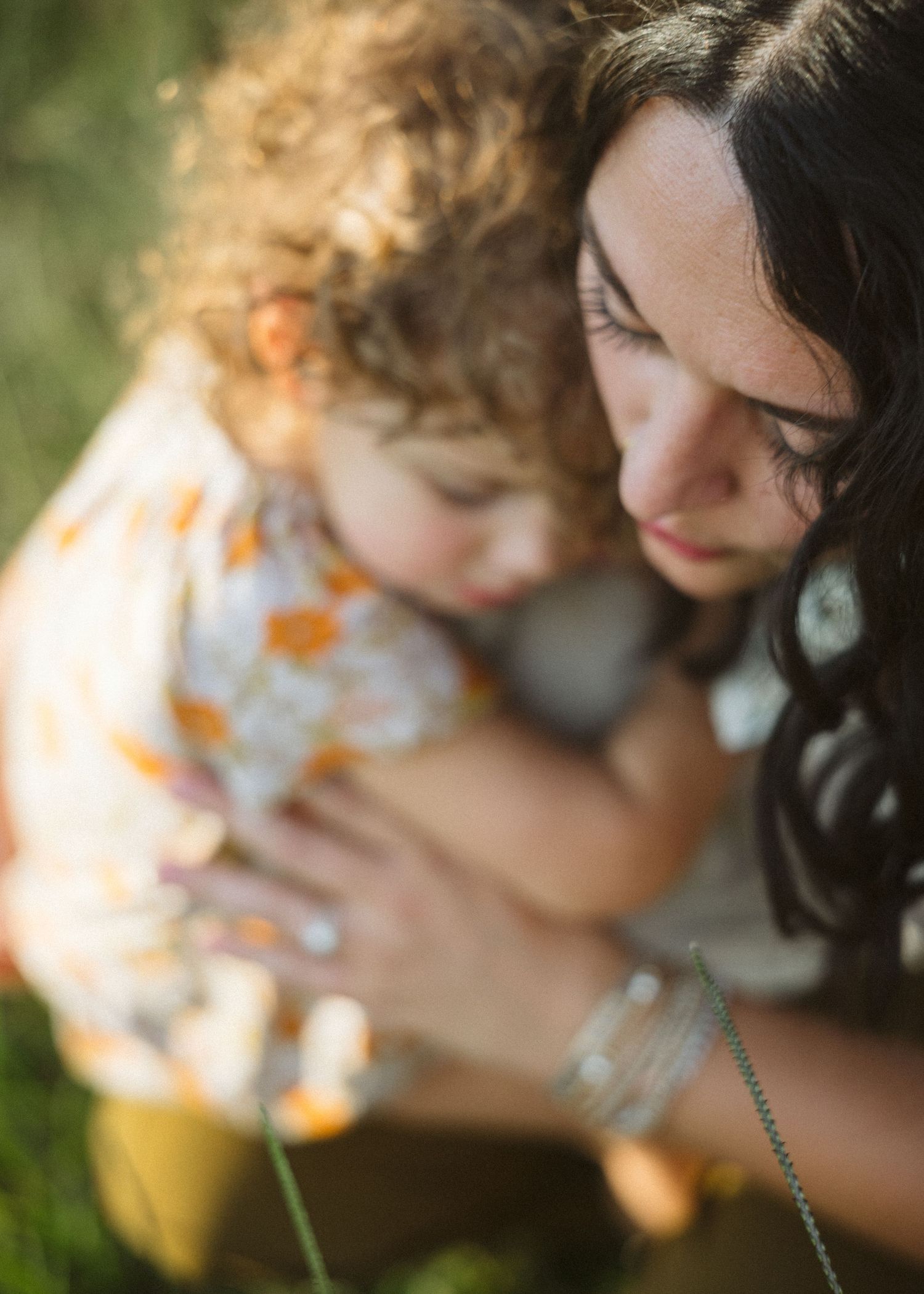 Dark-haired woman gently cradling a curly-haired toddler in a floral outfit while sitting in a sunlit grassy field.