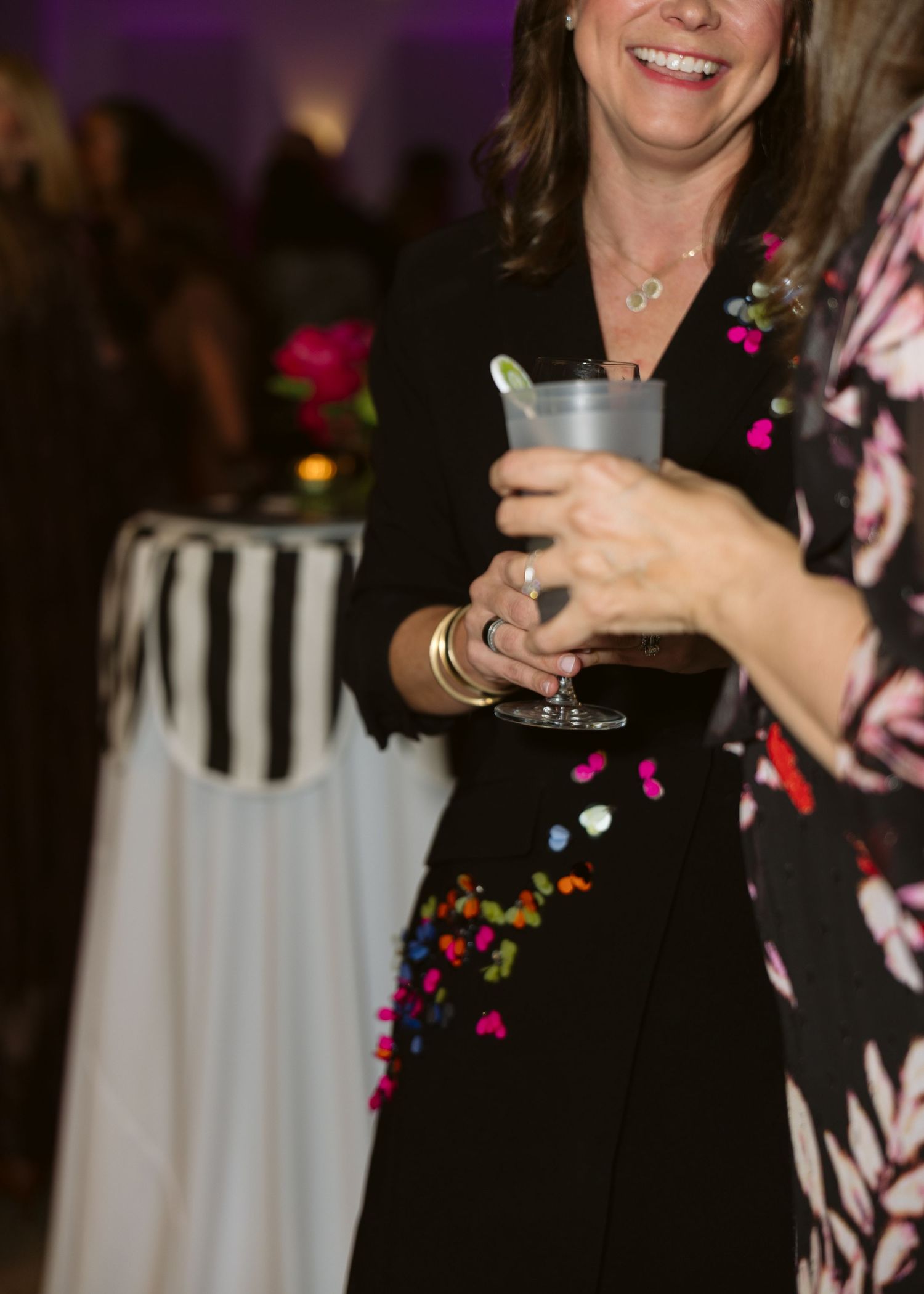 Smiling woman in a black dress with colorful confetti embellishments holds a drink at an evening party beside a striped table.