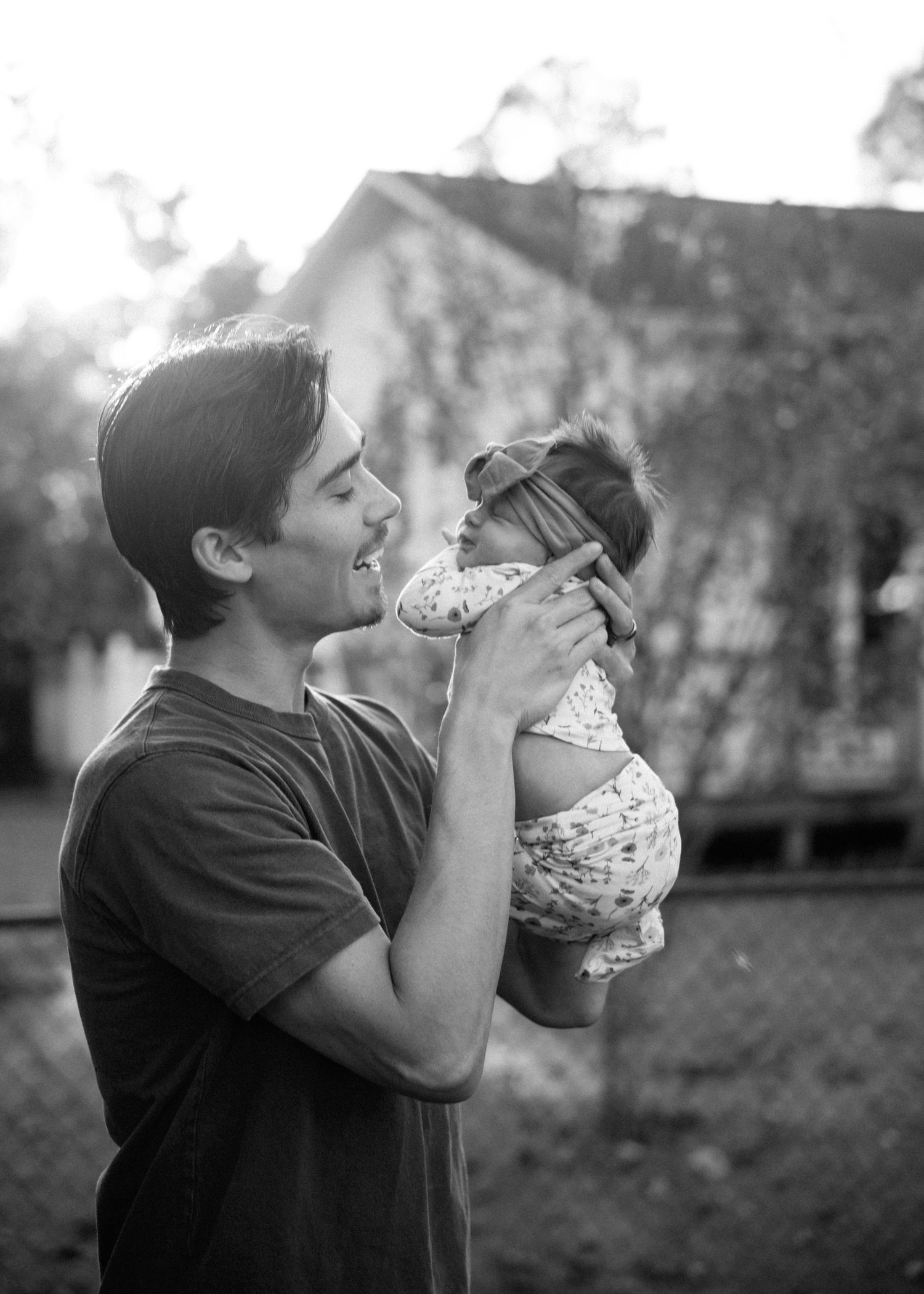 Smiling man in a t-shirt lifts a baby in a floral outfit and headband toward his face in a black and white outdoor portrait.