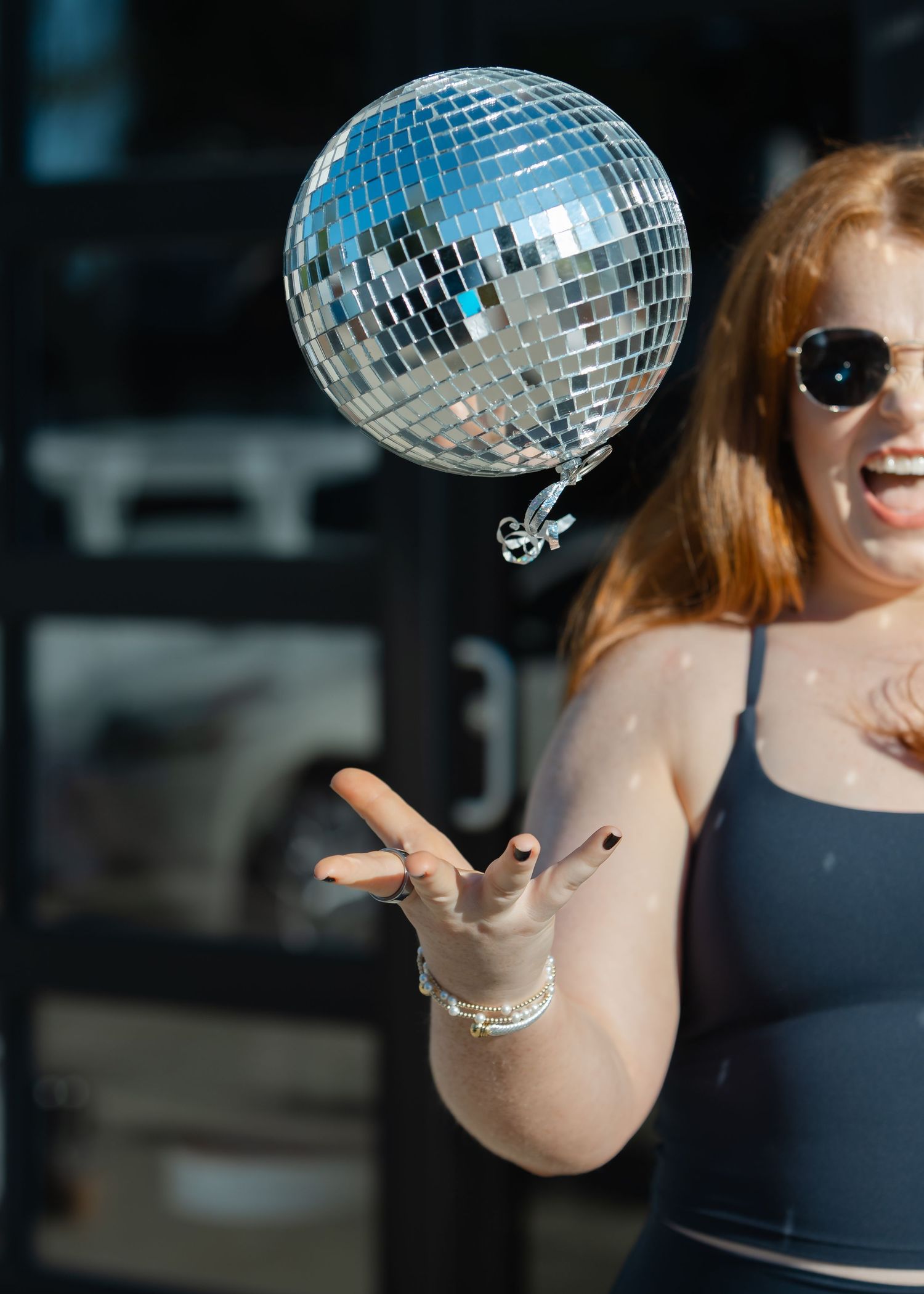 Woman with red hair tossing a mirrored disco ball in the air while wearing sunglasses outdoors.