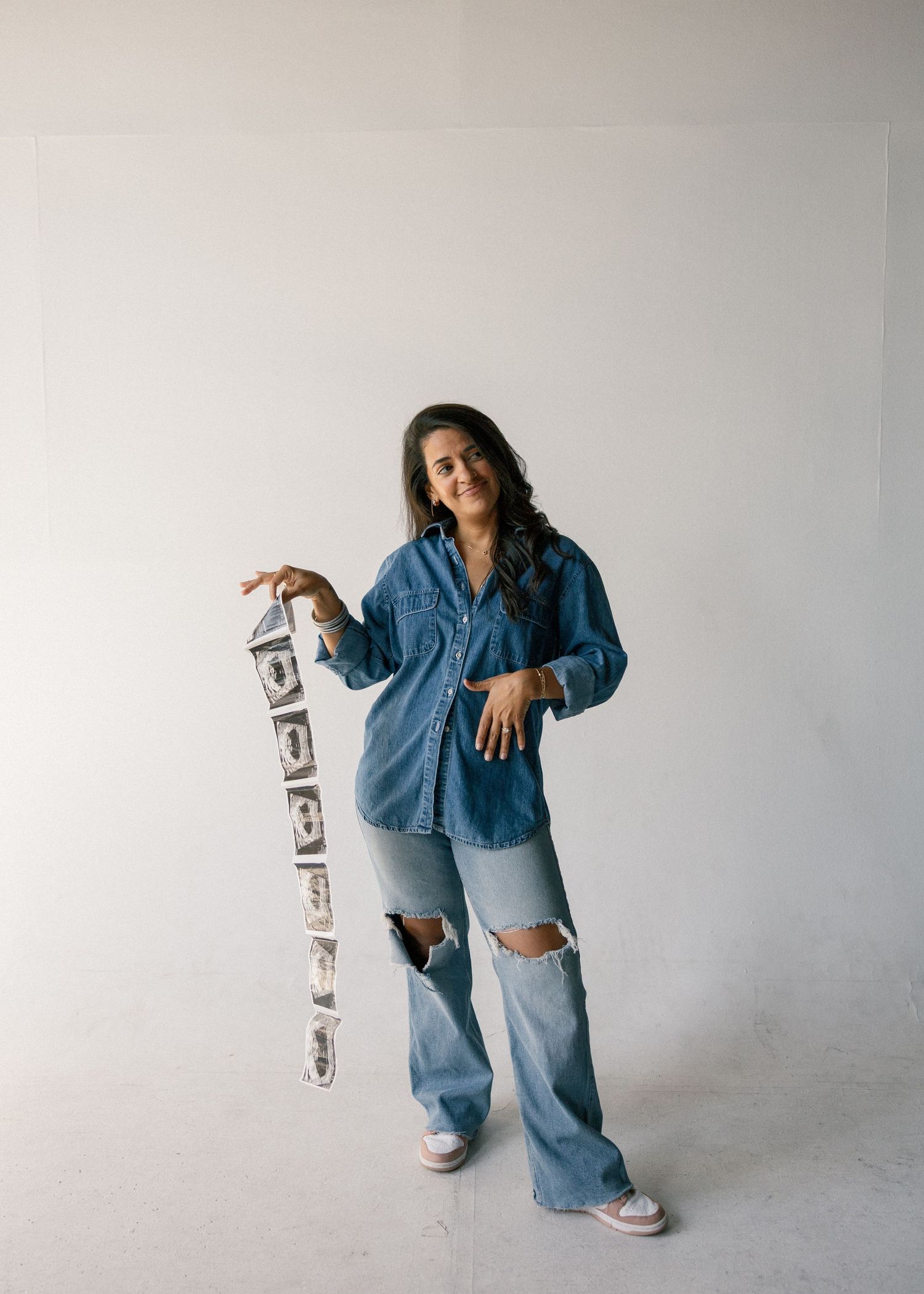 Woman in denim shirt and ripped jeans holds strip of photo booth pictures in studio.