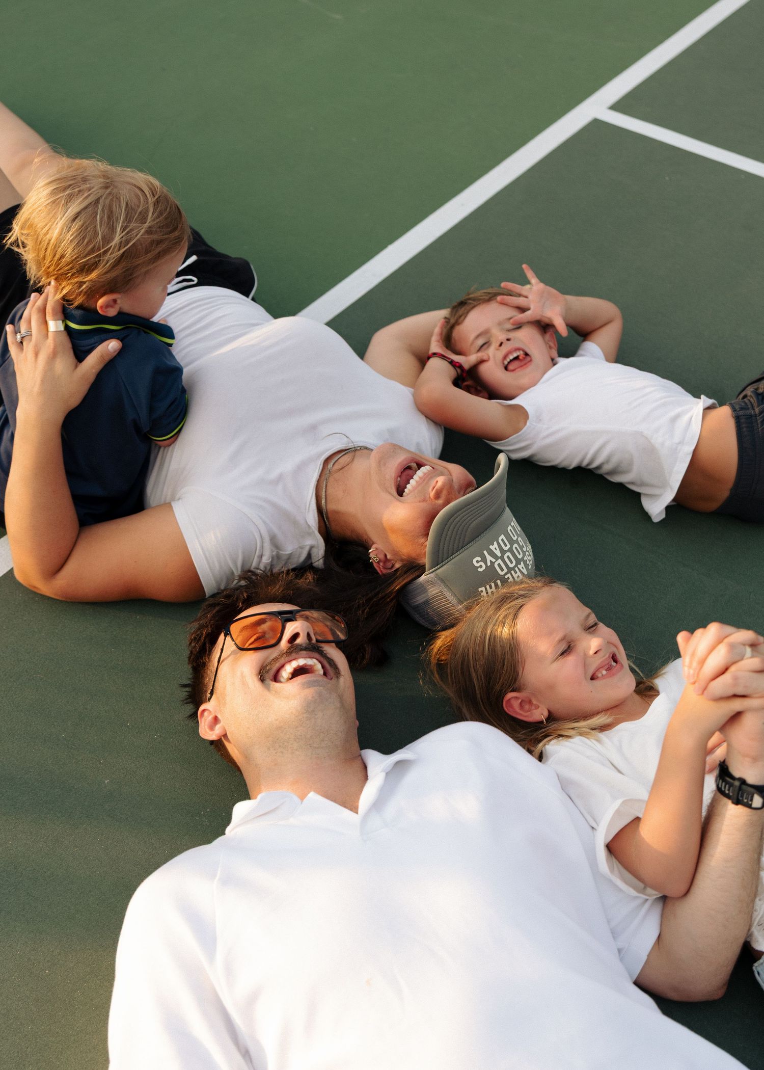 A family of five lies on a tennis court laughing together in the warm evening light.