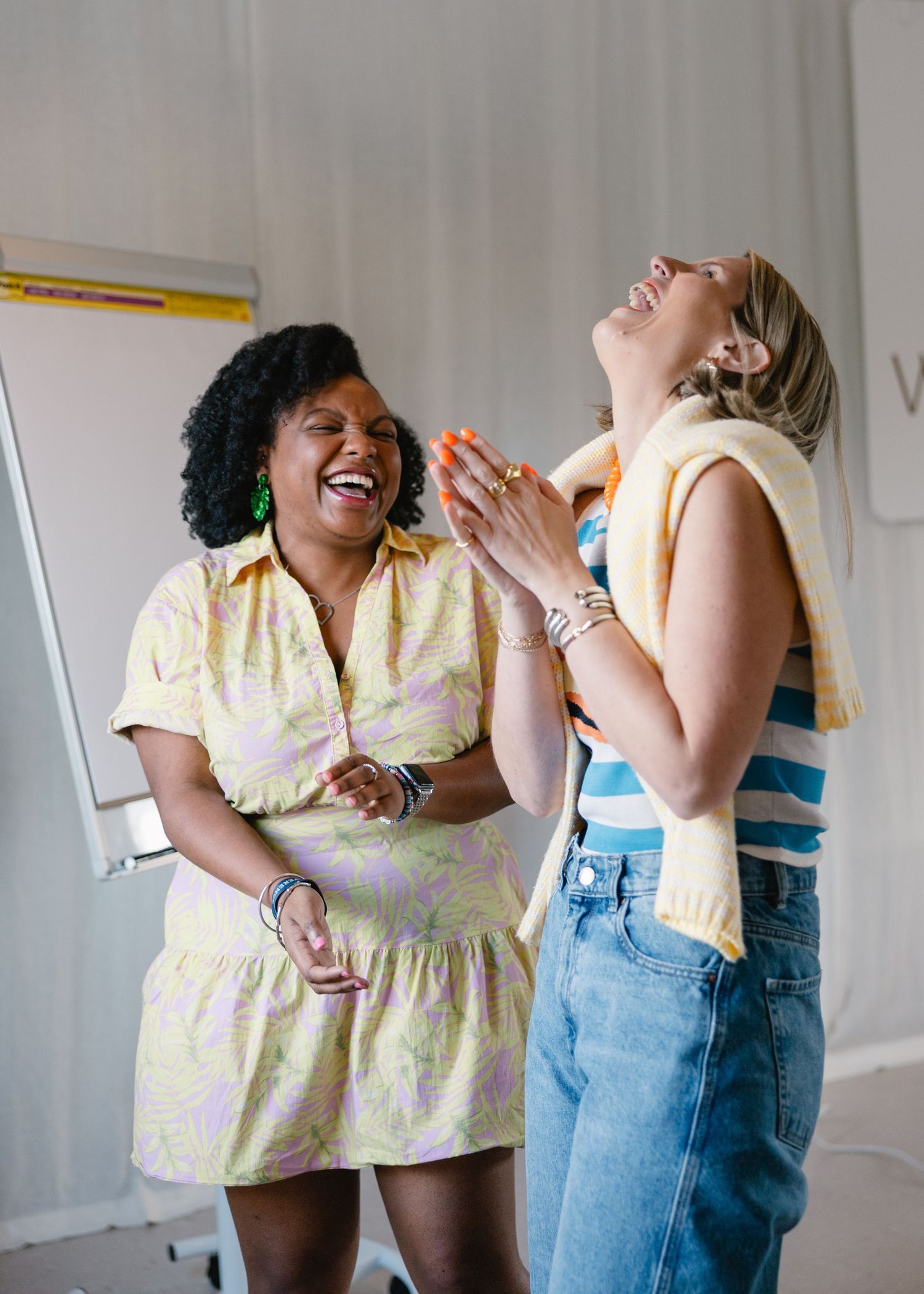 Two women laughing joyfully together in a bright room, one wearing a yellow floral dress and the other a striped top with jeans.
