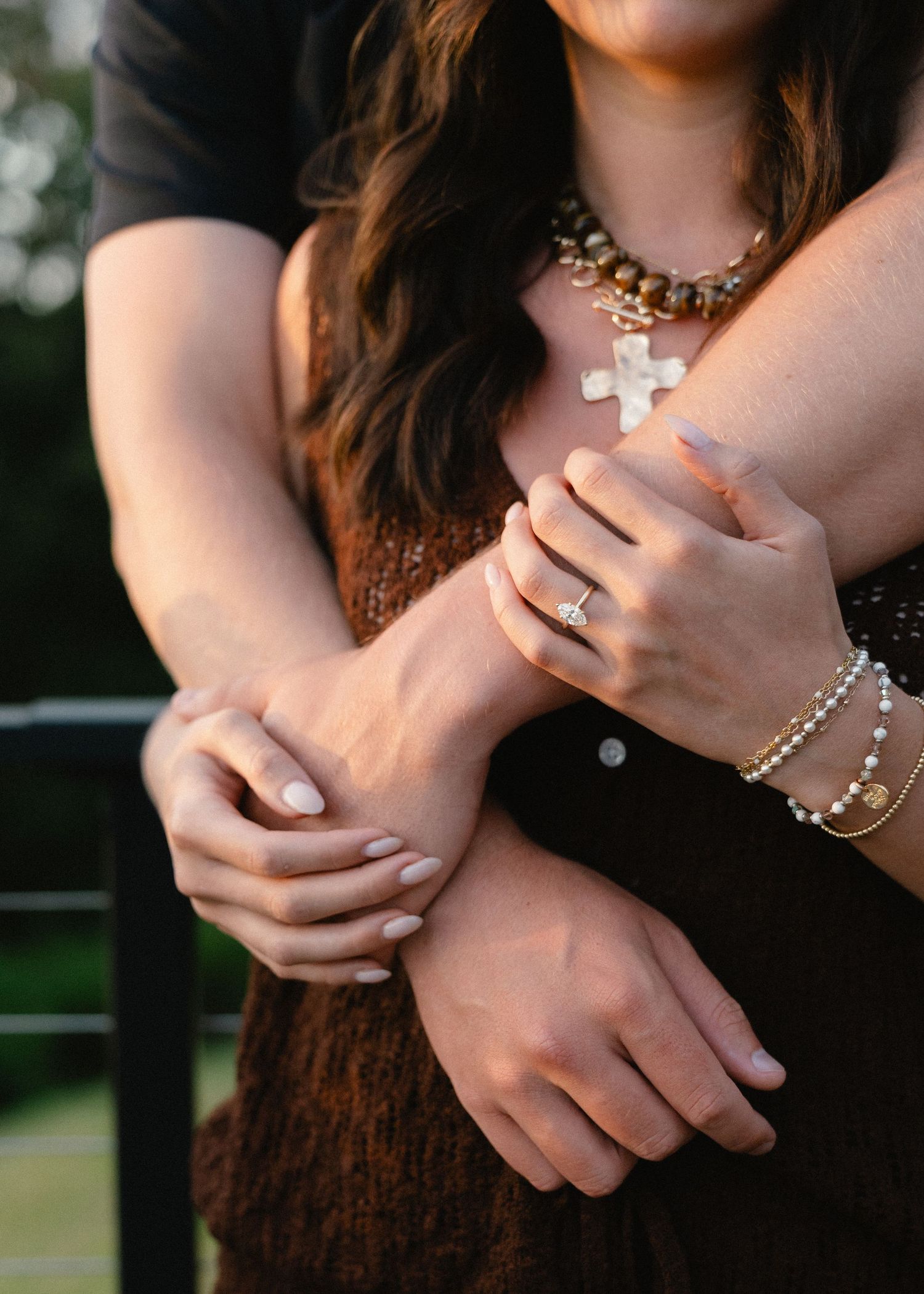 Couple embracing outdoors as a woman in a brown knit top wears a cross necklace, engagement ring and beaded bracelets.