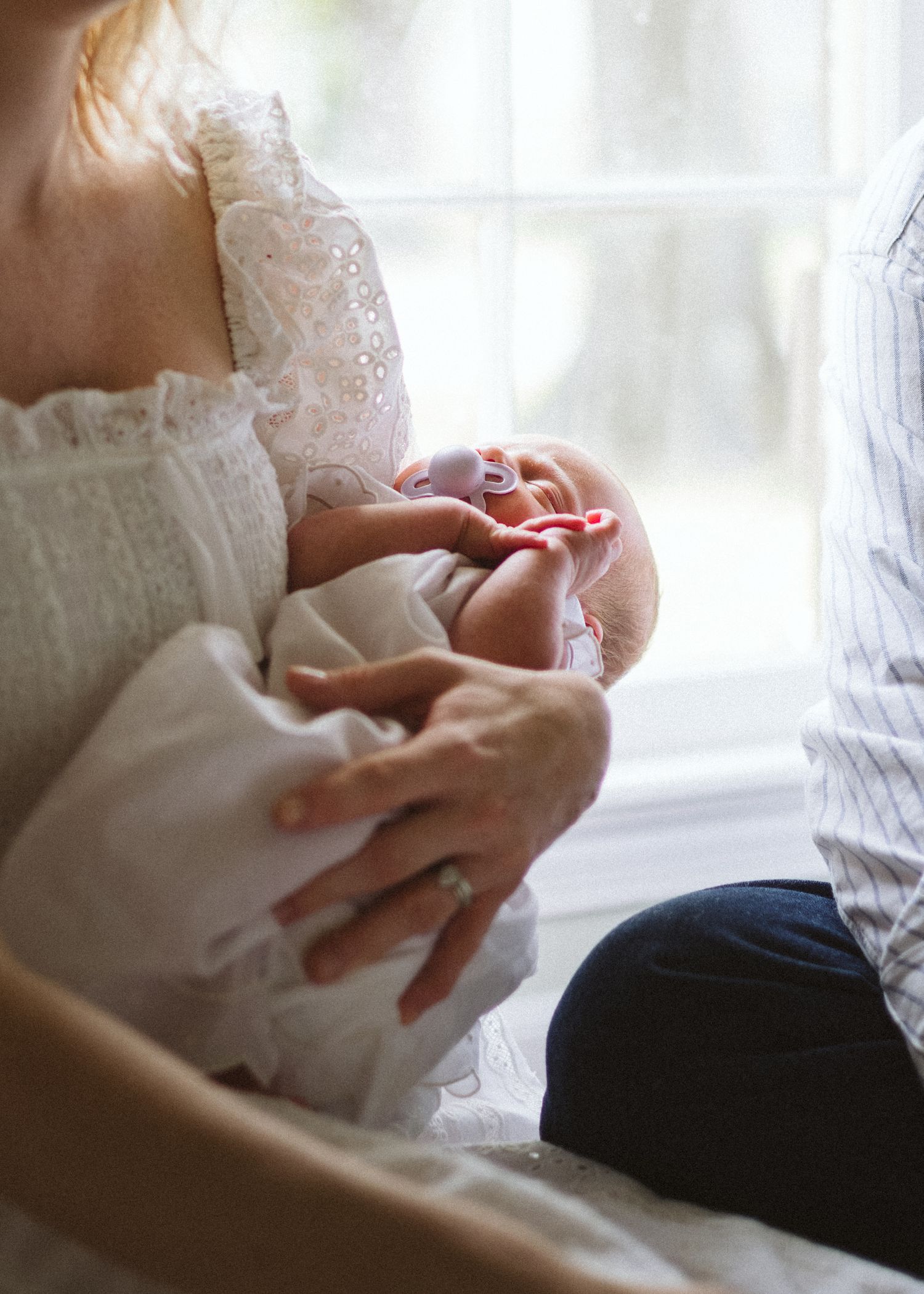 Mother in a white eyelet dress cradles a sleeping newborn baby wrapped in a soft blanket beside a sunlit window.
