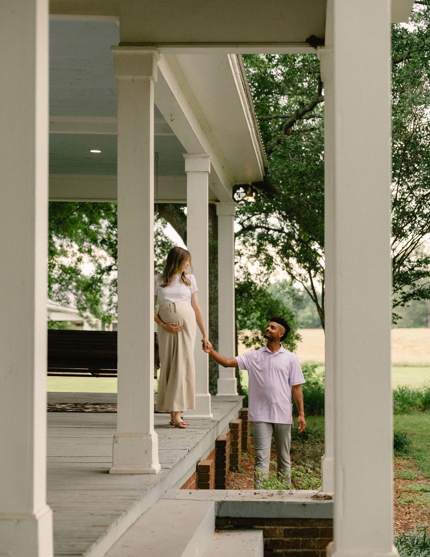 Pregnant woman stands on farmhouse porch as partner reaches up to her from the garden steps.