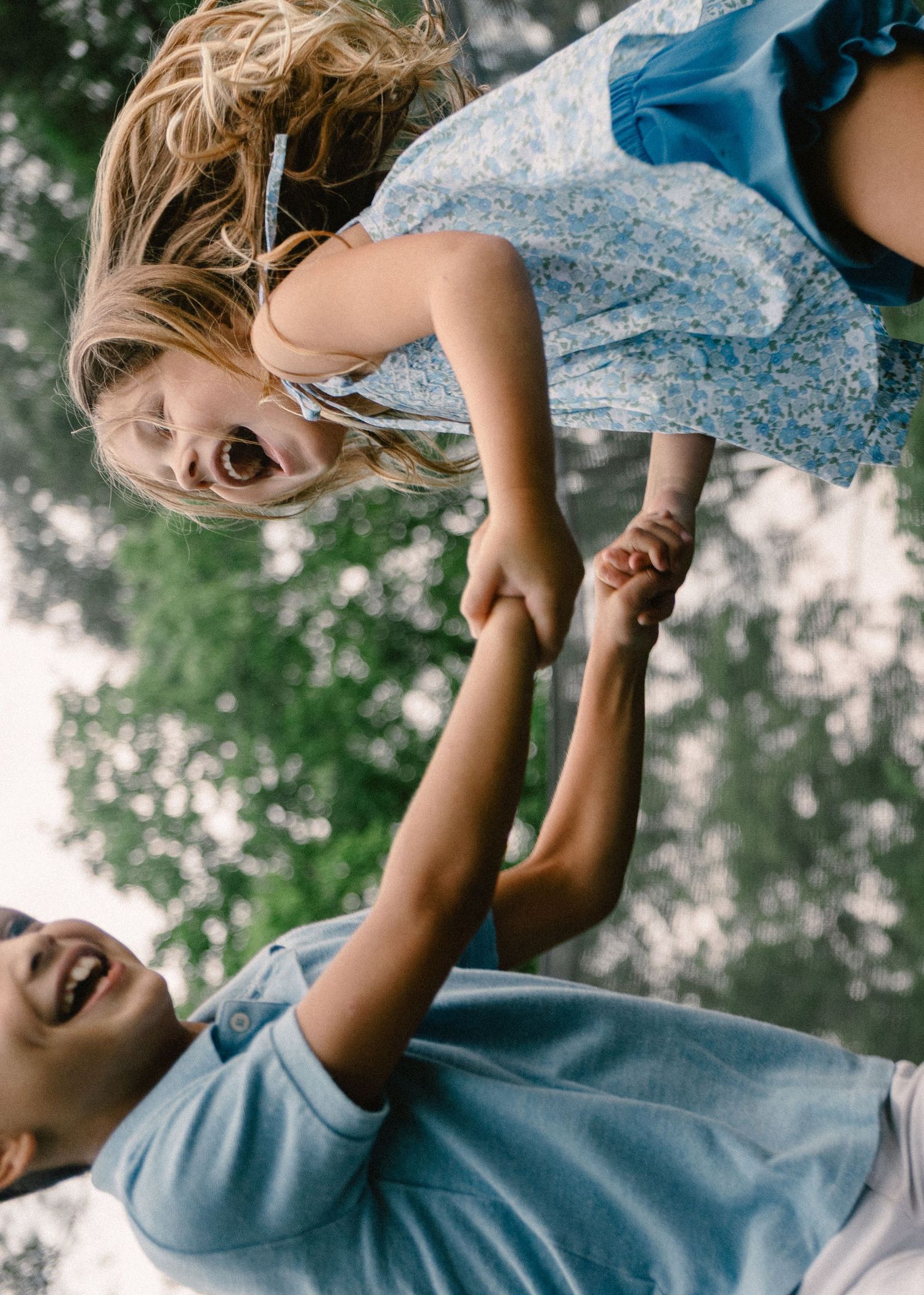 Father joyfully lifting laughing young girl in blue floral dress into the air outdoors.