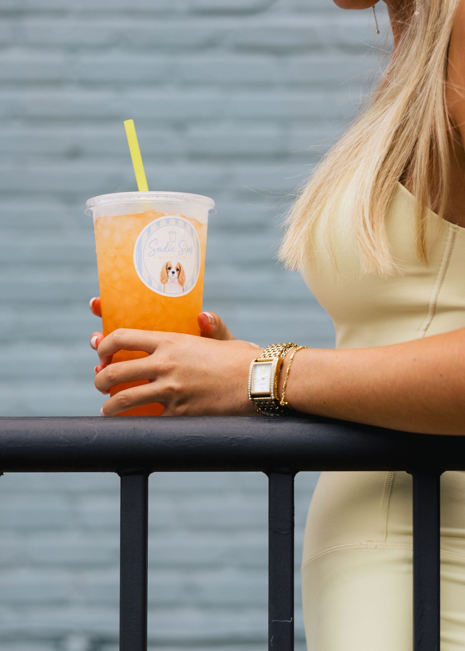 Blonde woman holding an orange iced drink with a Sadie Sips logo cup and yellow straw.