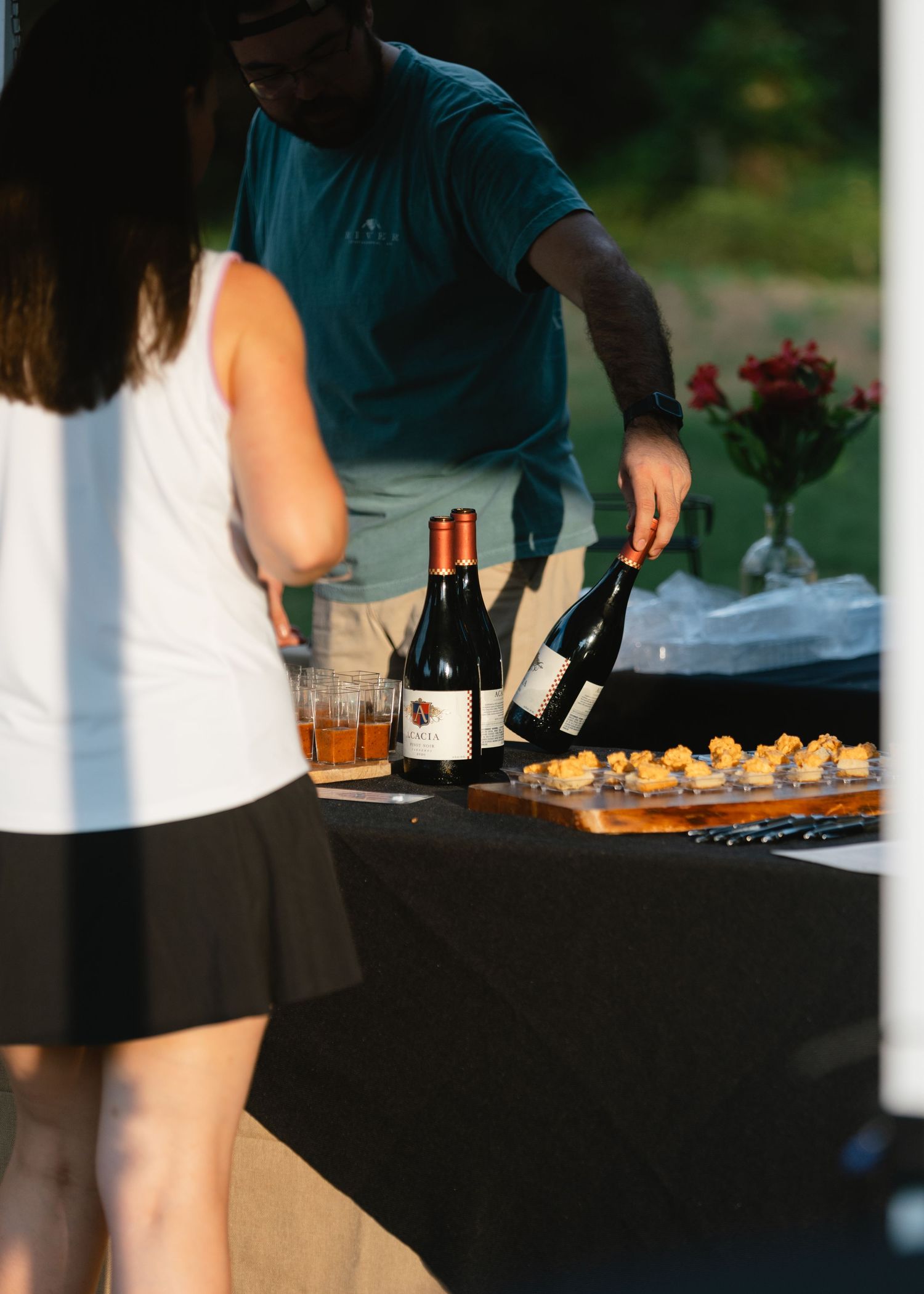 Man in a teal shirt pours red wine at an outdoor tasting table with wine bottles, appetizers, and a guest in a white tank top.