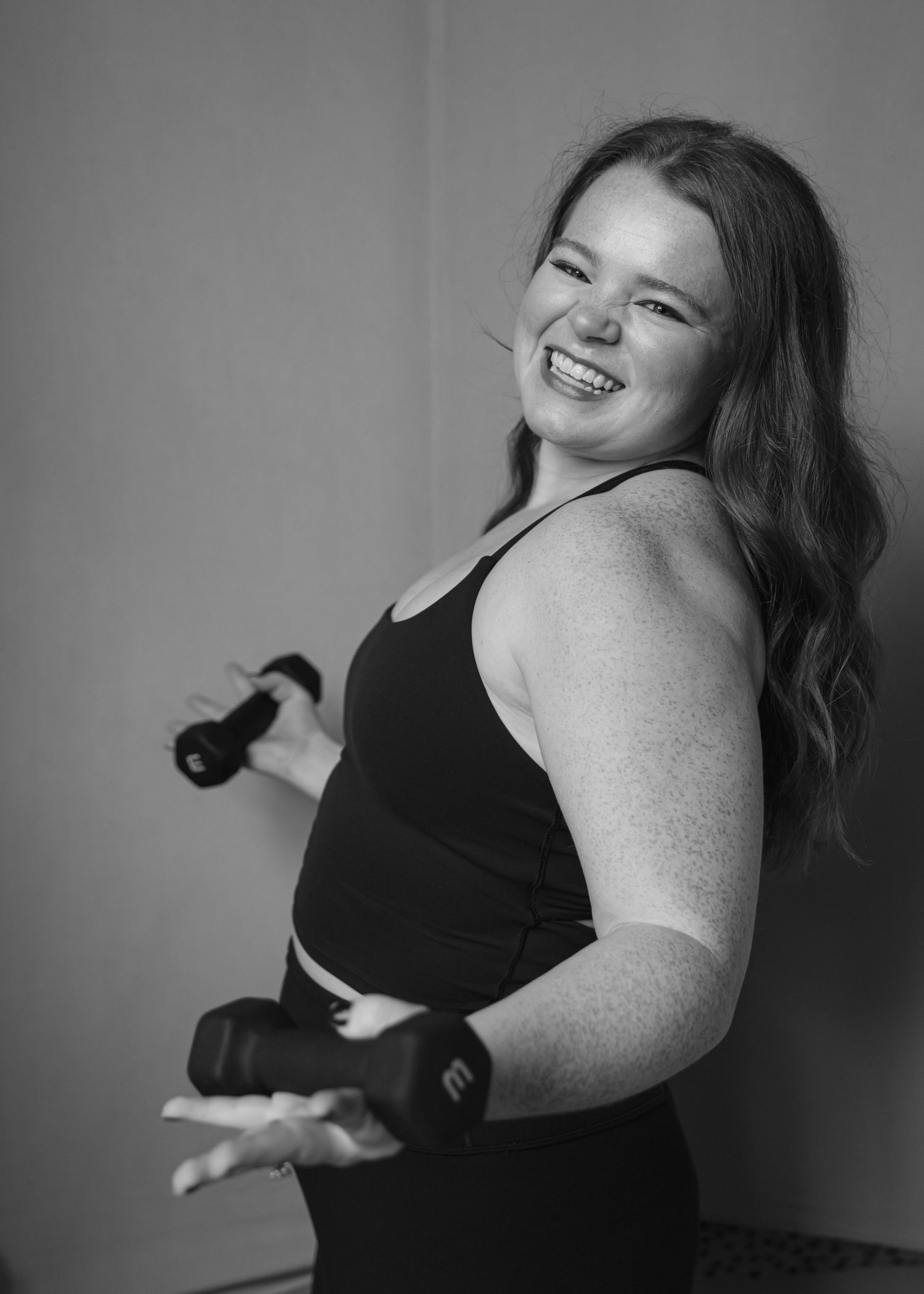 A smiling young woman in a black sports bra holds dumbbells in a black and white fitness photo.