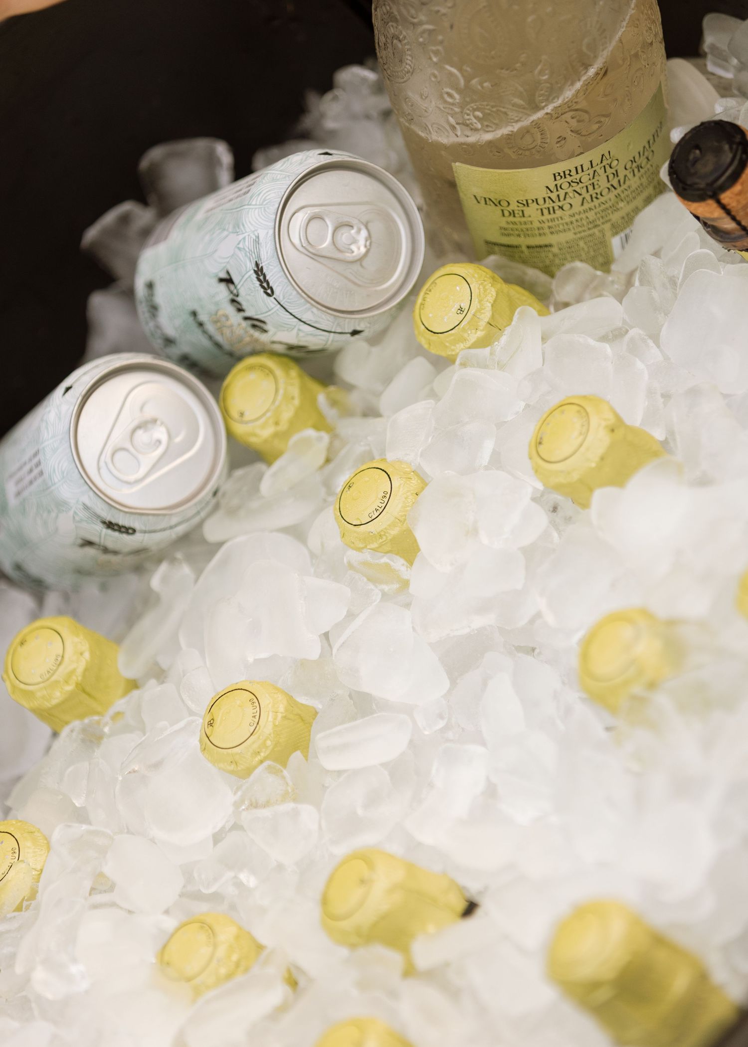 Gold-foiled sparkling wine bottles and white beverage cans chilling in a bed of crushed ice, ready to serve at a party.