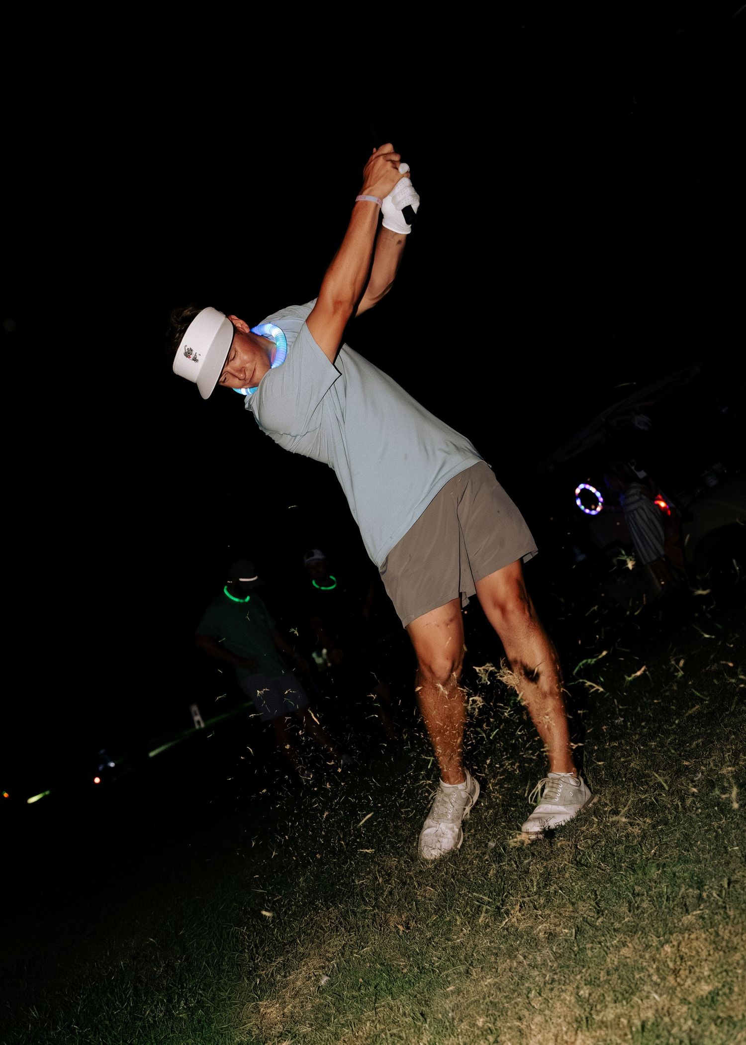 Man in white cap and grey polo completes a powerful golf swing at night outdoors.