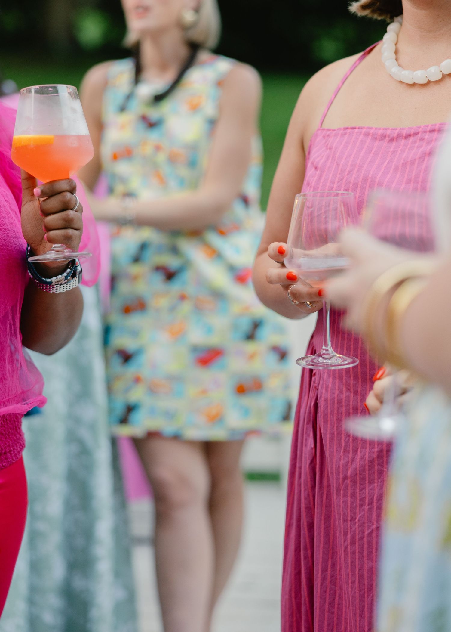 Women holding colorful cocktails and wine glasses in vibrant summer dresses at an outdoor garden party.
