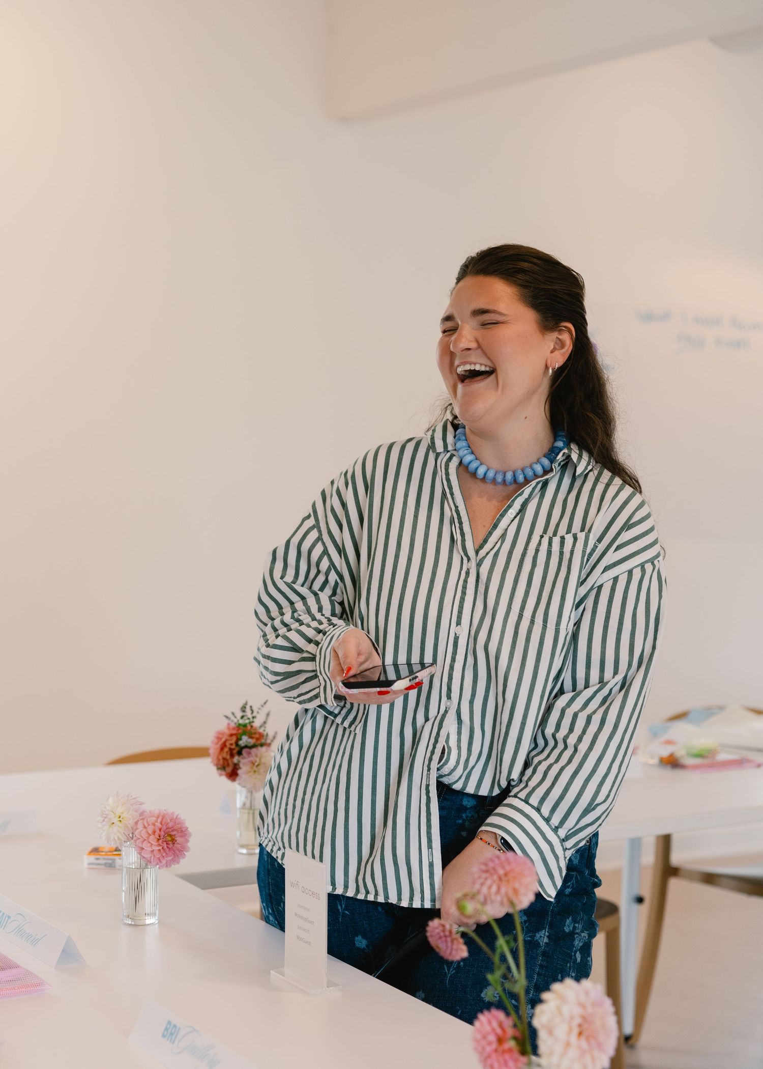 Woman in striped shirt laughing at a table with pink flowers in a white studio.